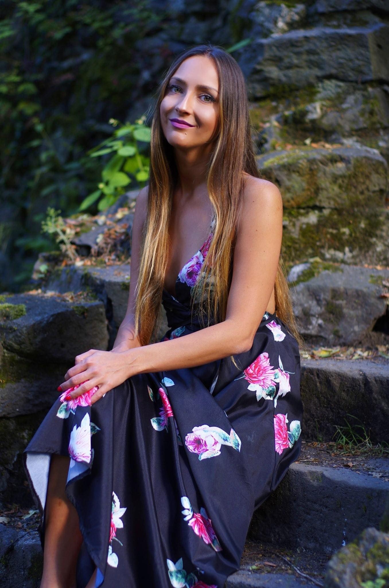 Portret młodej kobiety z długimi włosami w kwiecistej sukience, siedzącej na kamiennych schodach z mchem w tle, z delikatnym uśmiechem i pomalowanymi na czerwono paznokciami.