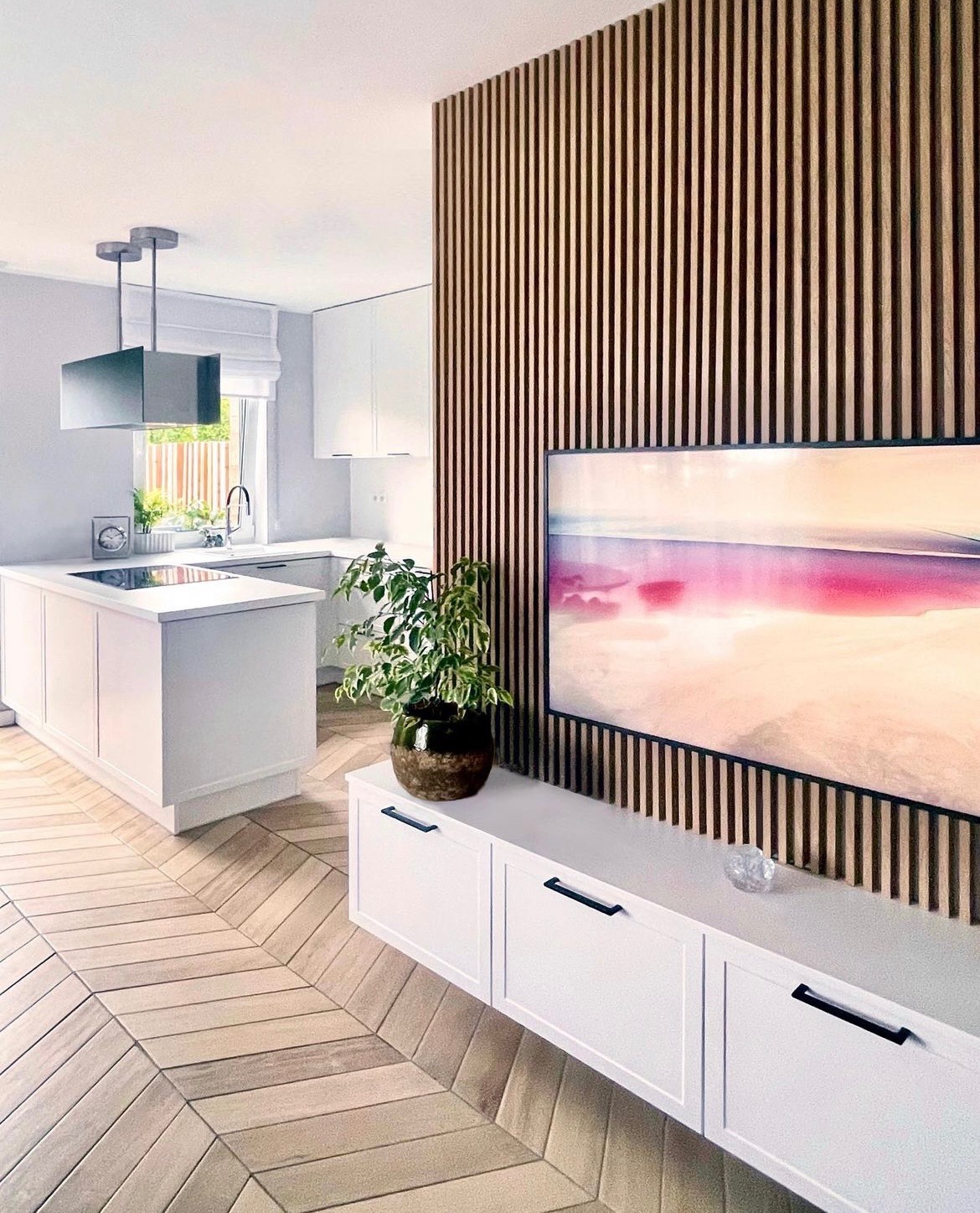 Nowoczesna kuchnia z wyspą, parkietem w jodełkę, ścianą z drewnianych lameli i białymi szafkami pod telewizorem.