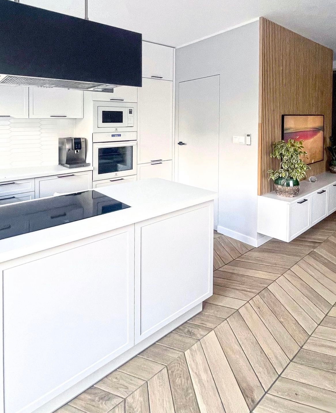 Jasna kuchnia z wyspą, drewnianą podłogą ułożoną we wzór jodełki i ścianą z drewnianych lameli, minimalistyczny design z czarnymi akcentami.