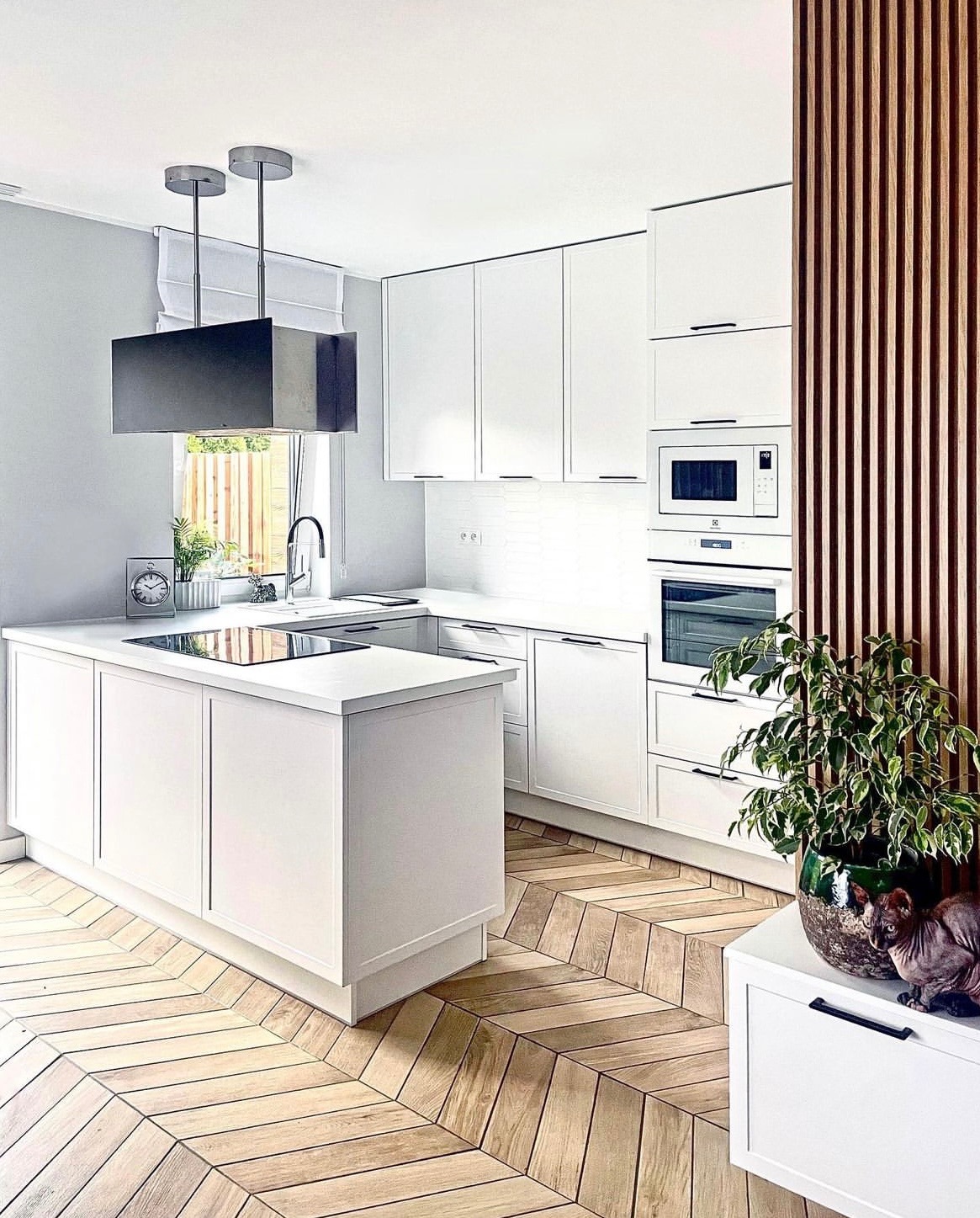 Jasna kuchnia z wyspą, białe szafki, parkiet ułożony w jodełkę, okap w formie prostopadłościanu zawieszony nad wyspą, drewniana ażurowa ścianka działowa, kot sfinks siedzący na szafce.