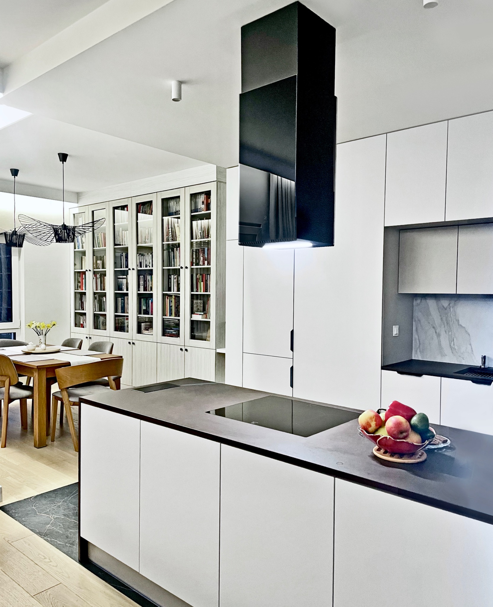 Nowoczesna kuchnia z wyspą i czarnym okapem, widok na stół jadalny z biblioteczką w tle.