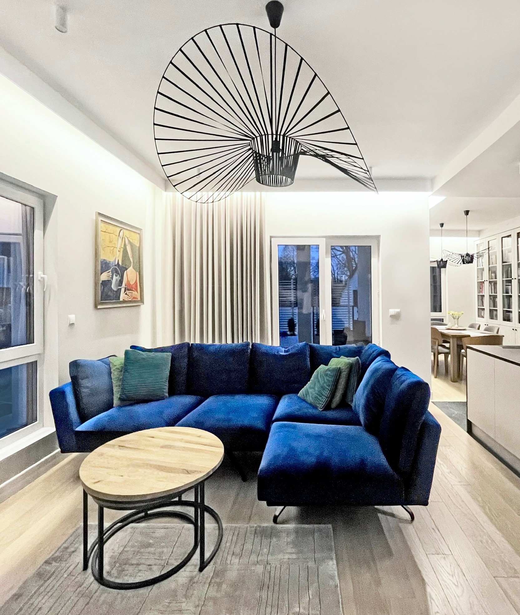 Salon z narożną, welurową sofą w odcieniach niebieskiego, okrągłym stolikiem kawowym z drewnianym blatem i metalową podstawą oraz unikalną, wiszącą lampą z metalowych prętów.