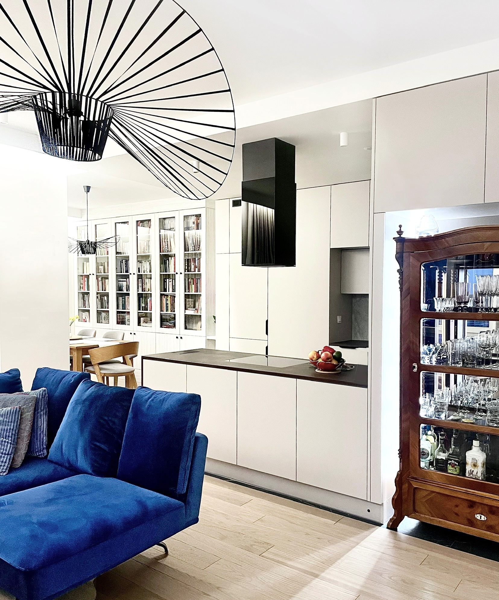 Nowoczesna kuchnia z białymi szafkami, czarnym okapem i wyspą z owocami, widok na salon z niebieską sofą i biblioteczką za stołem jadalnym, oświetlona designerską lampą.