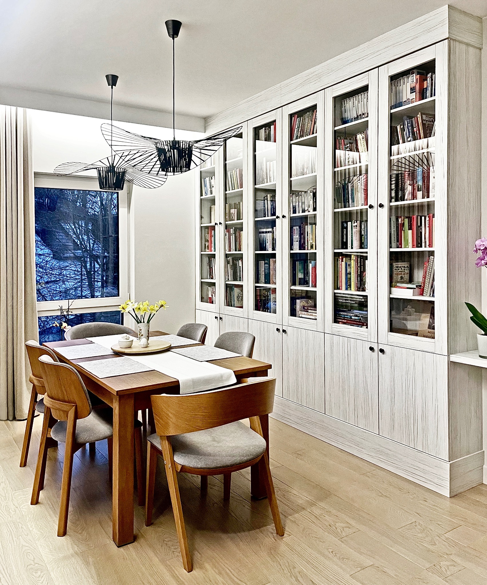 Jadalnia z drewnianym stołem, szarymi krzesłami i zabudowaną biblioteczką z przeszklonymi drzwiami, oświetlona wiszącymi lampami w kształcie kapeluszy.