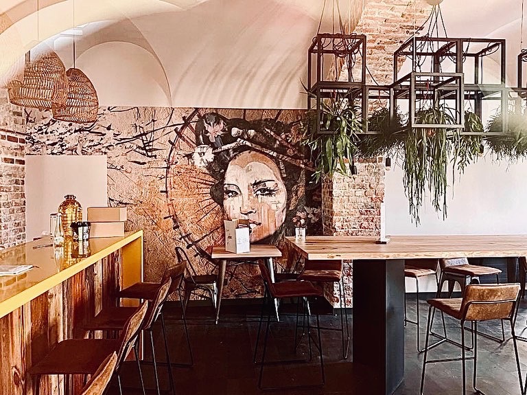 Wnętrze kawiarni z muralem kobiety na ceglanej ścianie, drewnianym barem z żółtym blatem, wiszącymi lampami z wikliny i metalowymi konstrukcjami z roślinami.