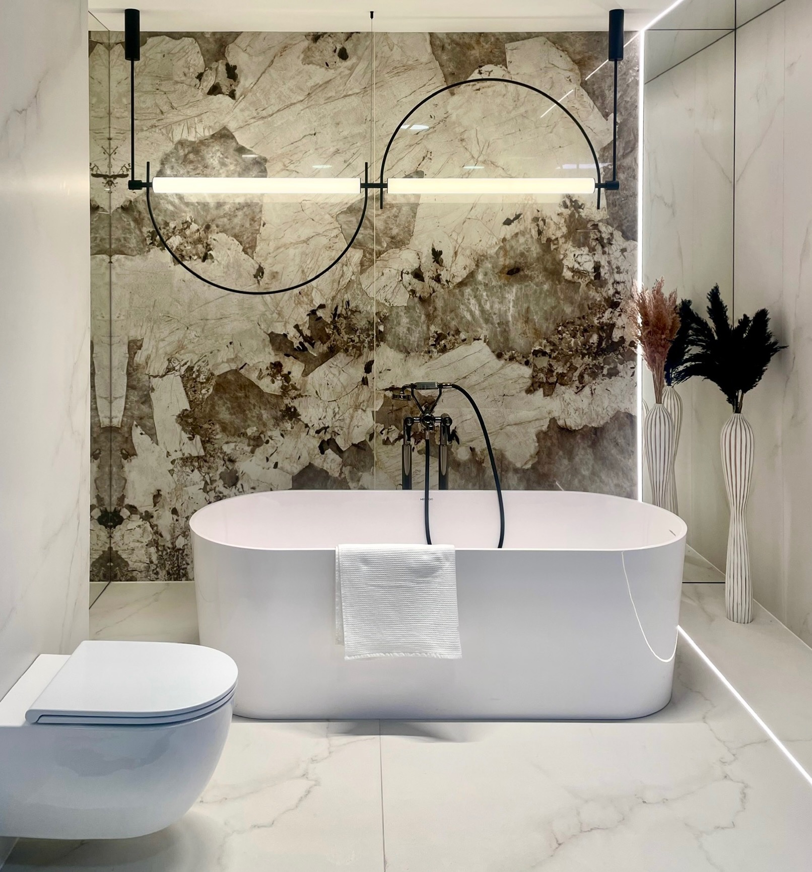 Nowoczesna łazienka we Wrocławiu z wolnostojącą białą wanną, wiszącą toaletą i marmurową ścianą akcentującą, oświetlona designerską lampą z czarnymi półokręgami i białym światłem.