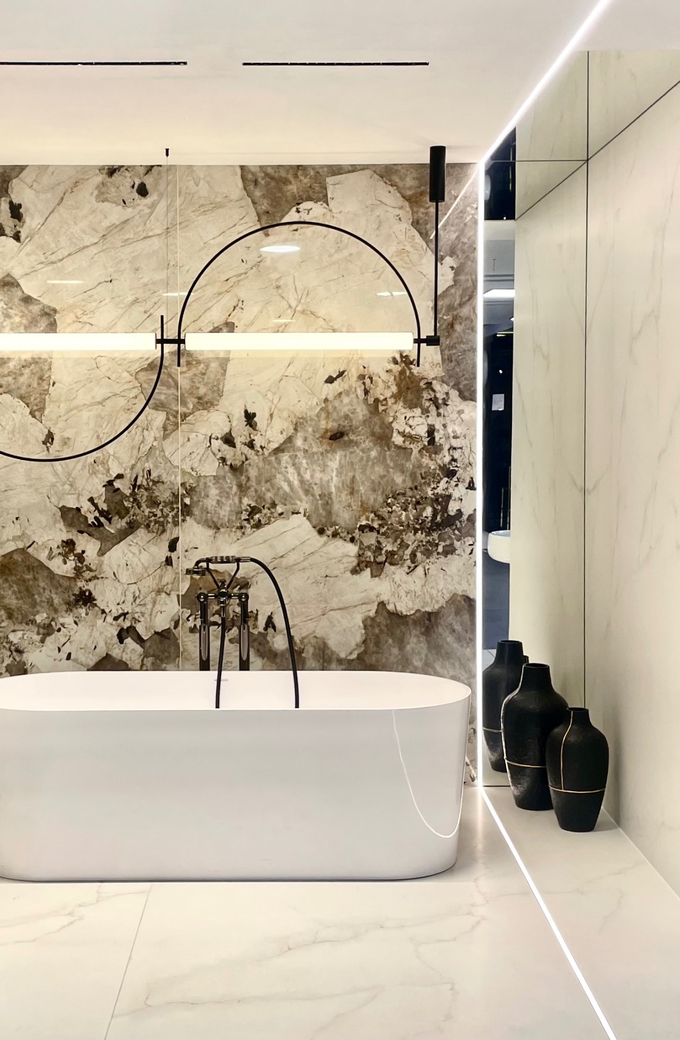 Nowoczesna łazienka z wolnostojącą białą wanną, armaturą w stylu retro, marmurową ścianą akcentową i dekoracyjnym oświetleniem w kształcie półkola oraz czarnymi wazonami.