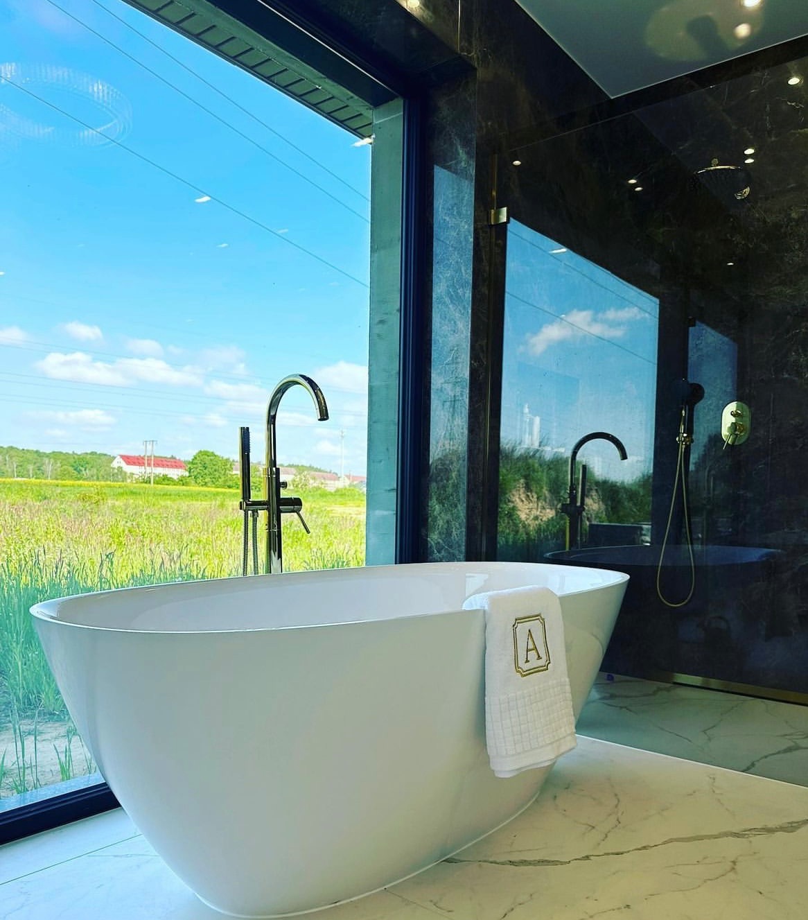 Nowoczesna łazienka z wolnostojącą białą wanną na marmurowej podłodze, widok na zielone pole przez duże okno, ręcznik z monogramem 'A'.
