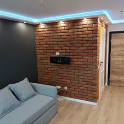Pokój dzienny po remoncie: ściana z cegły dekoracyjnej, szara ściana, sofa z poduszkami, oświetlenie LED sufitowe.