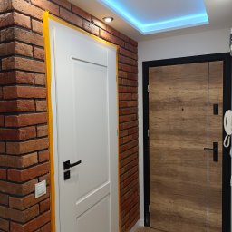 Cristal - Wnętrze mieszkania z dwiema drzwiami: białe drzwi z czarną klamką obok ściany z cegły, zabezpieczone żółtą taśmą malarską, oraz brązowe drzwi z czarną ramą, widoczne oświetlenie LED na suficie.