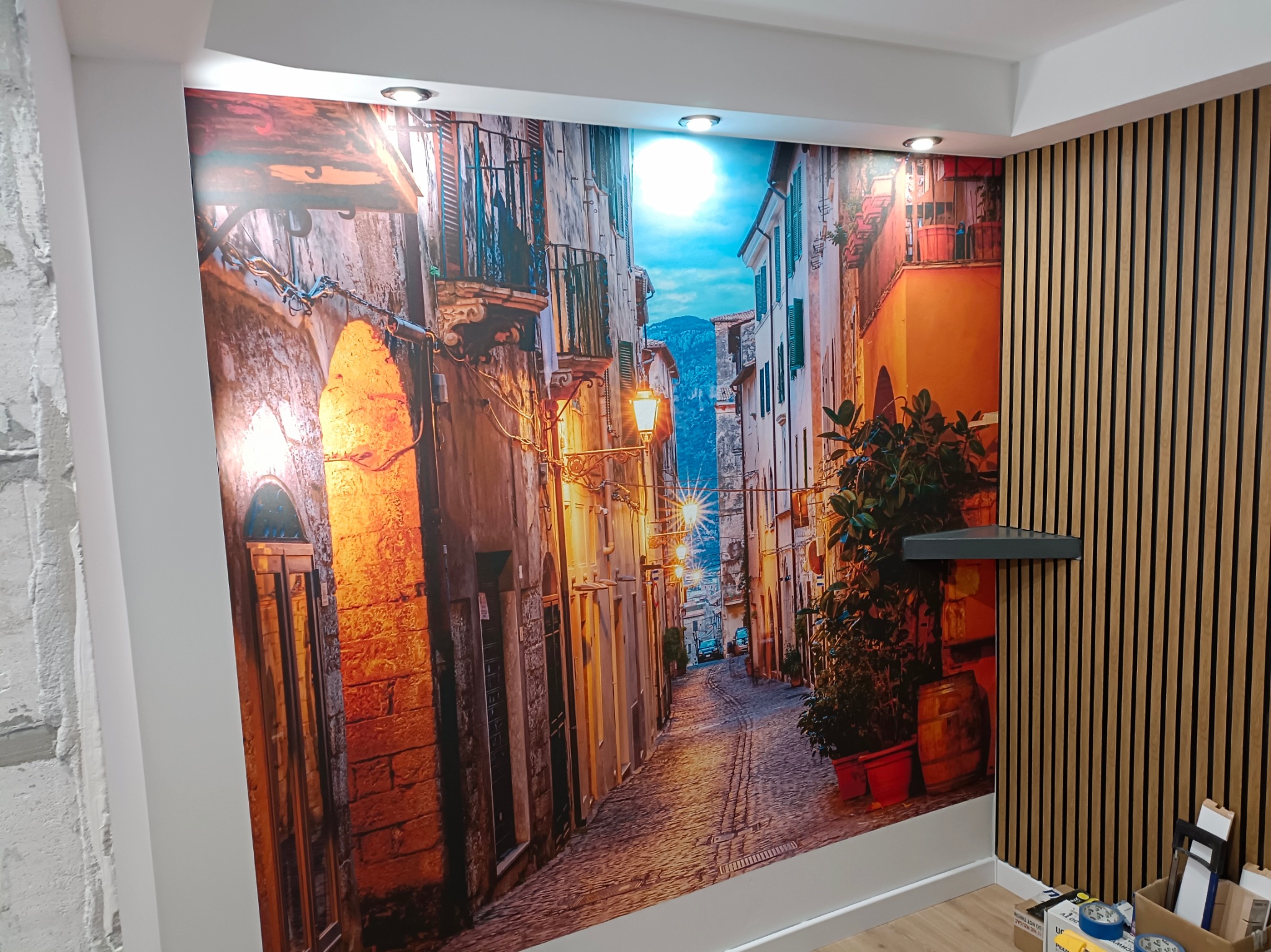 Fototapeta z motywem włoskiej uliczki w ciepłych barwach, połączona z drewnianą boazerią lamelową i fragmentem surowej ściany, oświetlona punktowymi lampami sufitowymi.