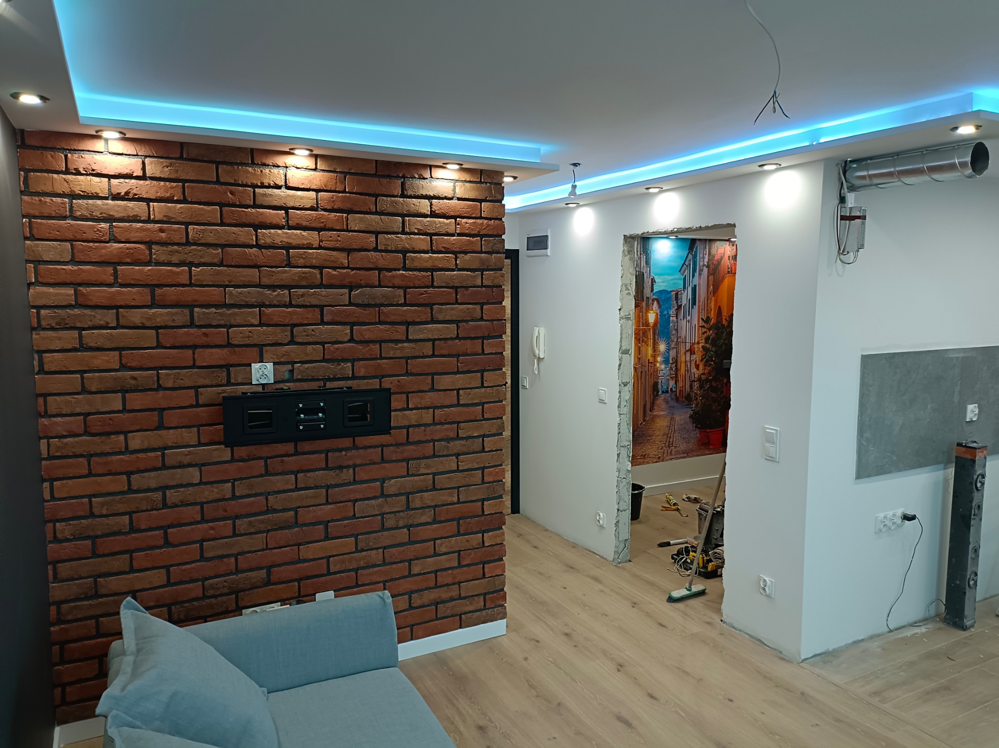 Salon w trakcie remontu: ściana z cegły dekoracyjnej z uchwytem na telewizor, sufit podwieszany z niebieskim oświetleniem LED, widoczne narzędzia i materiały remontowe w przejściu do innego...