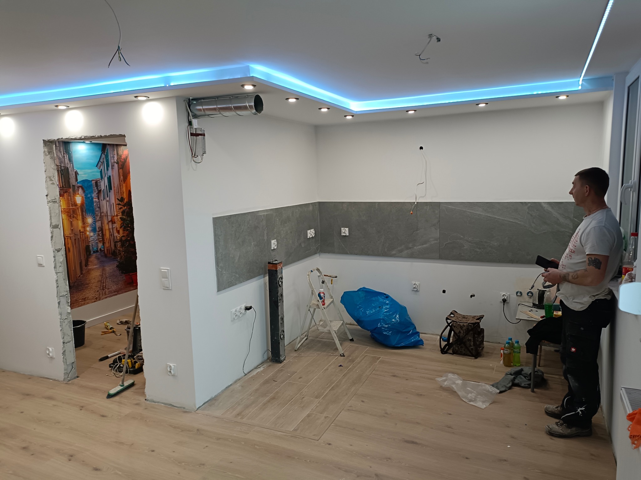 Wnętrze w trakcie remontu: położone szare płytki, drewnopodobne panele na podłodze, podwieszany sufit z niebieskim oświetleniem LED, widoczne narzędzia i materiały budowlane, pracownik z telefonem...