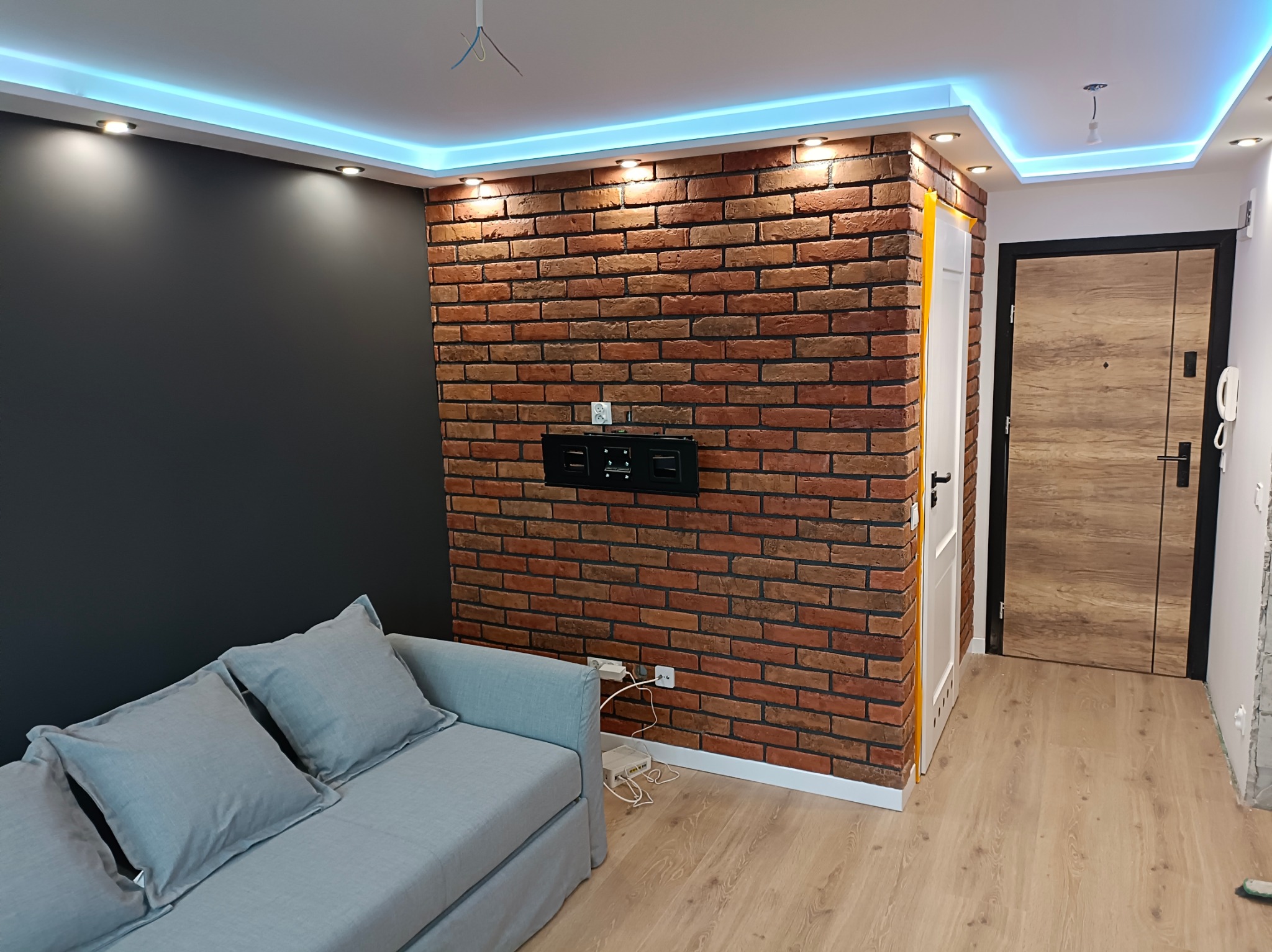 Pokój dzienny po remoncie: ściana z cegły dekoracyjnej, szara ściana, sofa z poduszkami, oświetlenie LED sufitowe.