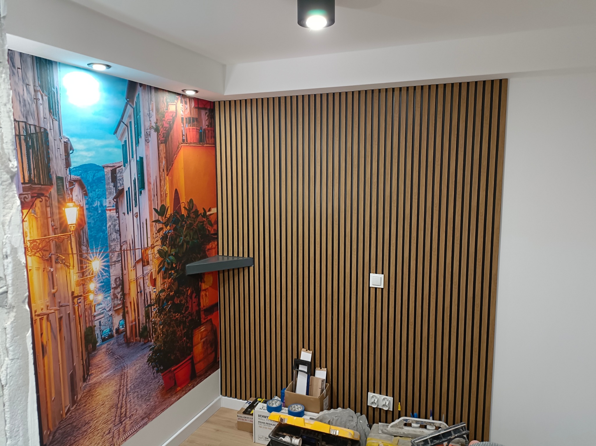 Pokój po remoncie: ściana z drewnianych paneli pionowych sąsiaduje z fototapetą przedstawiającą włoską uliczkę, widoczne narzędzia i materiały budowlane na podłodze.