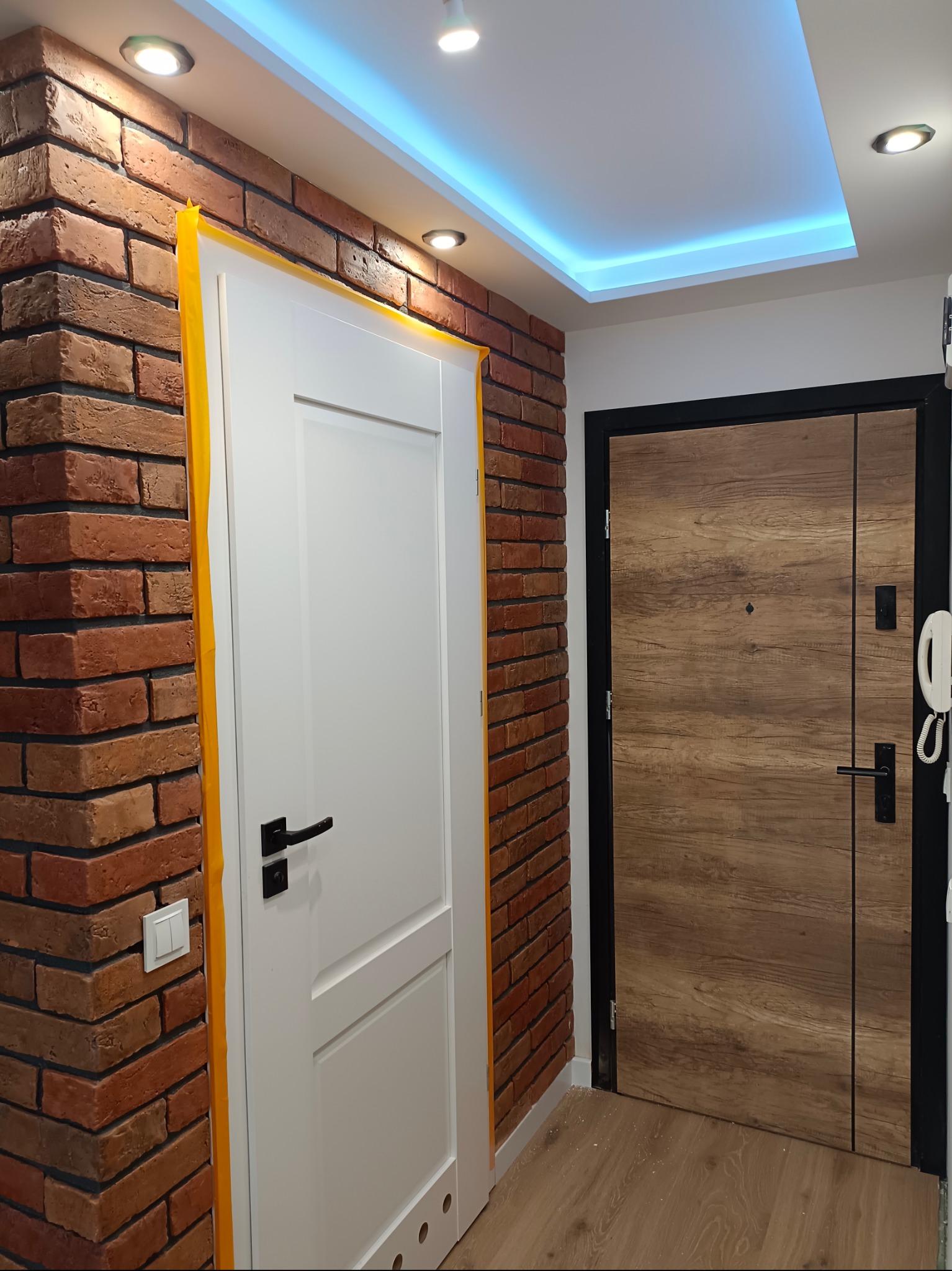 Wnętrze mieszkania z dwiema drzwiami: białe drzwi z czarną klamką obok ściany z cegły, zabezpieczone żółtą taśmą malarską, oraz brązowe drzwi z czarną ramą, widoczne oświetlenie LED na suficie.