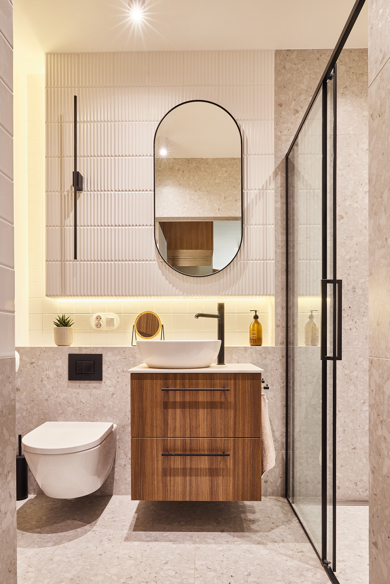 Nowoczesna łazienka z wiszącą toaletą, szafką z umywalką i kabiną prysznicową. Beżowe płytki z terazzo, oświetlenie LED i czarne akcenty.