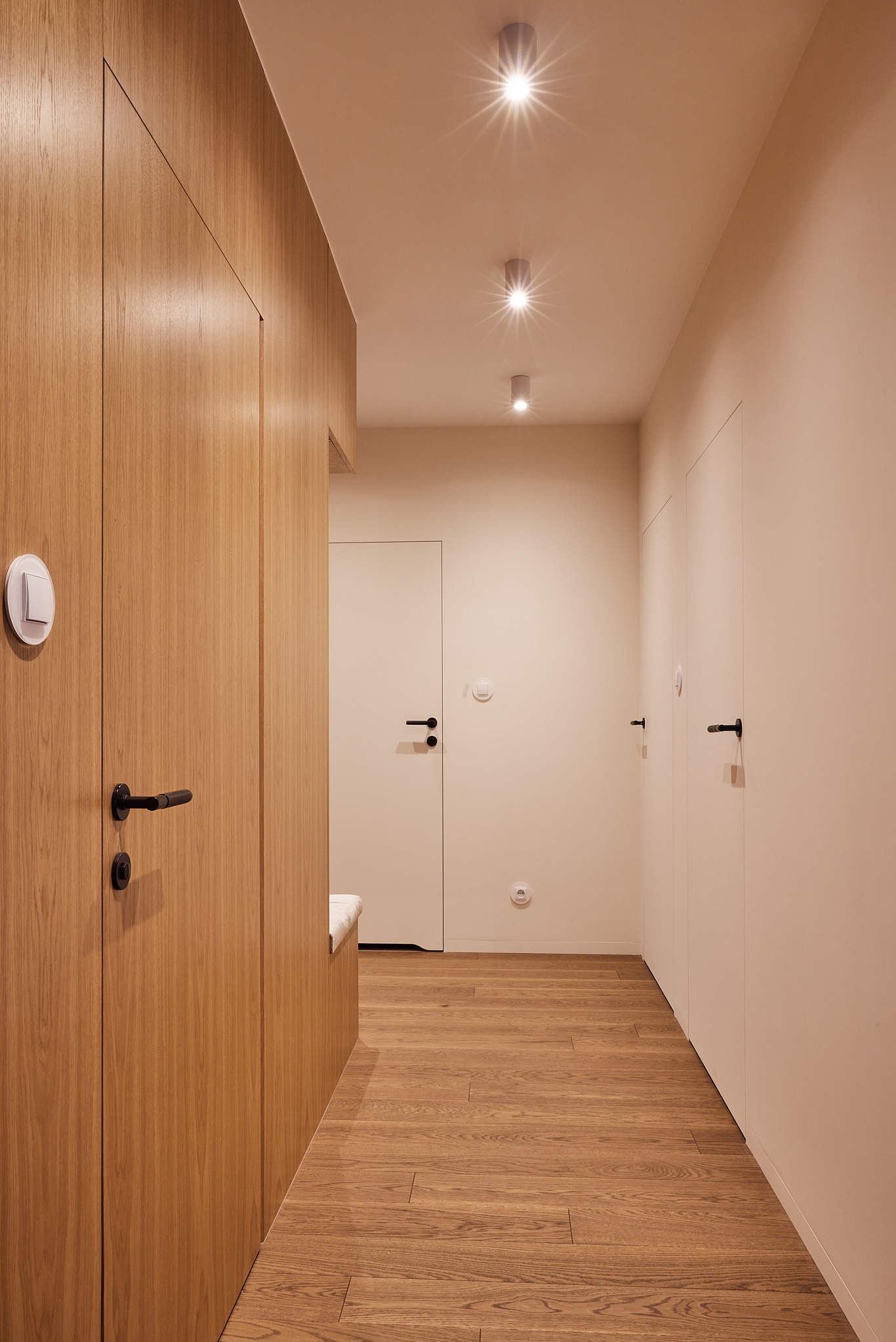 Nowoczesny korytarz z drewnianą podłogą i zabudową, minimalistyczne białe drzwi z czarnymi klamkami, oświetlenie punktowe. Jasna kolorystyka, spójny design.