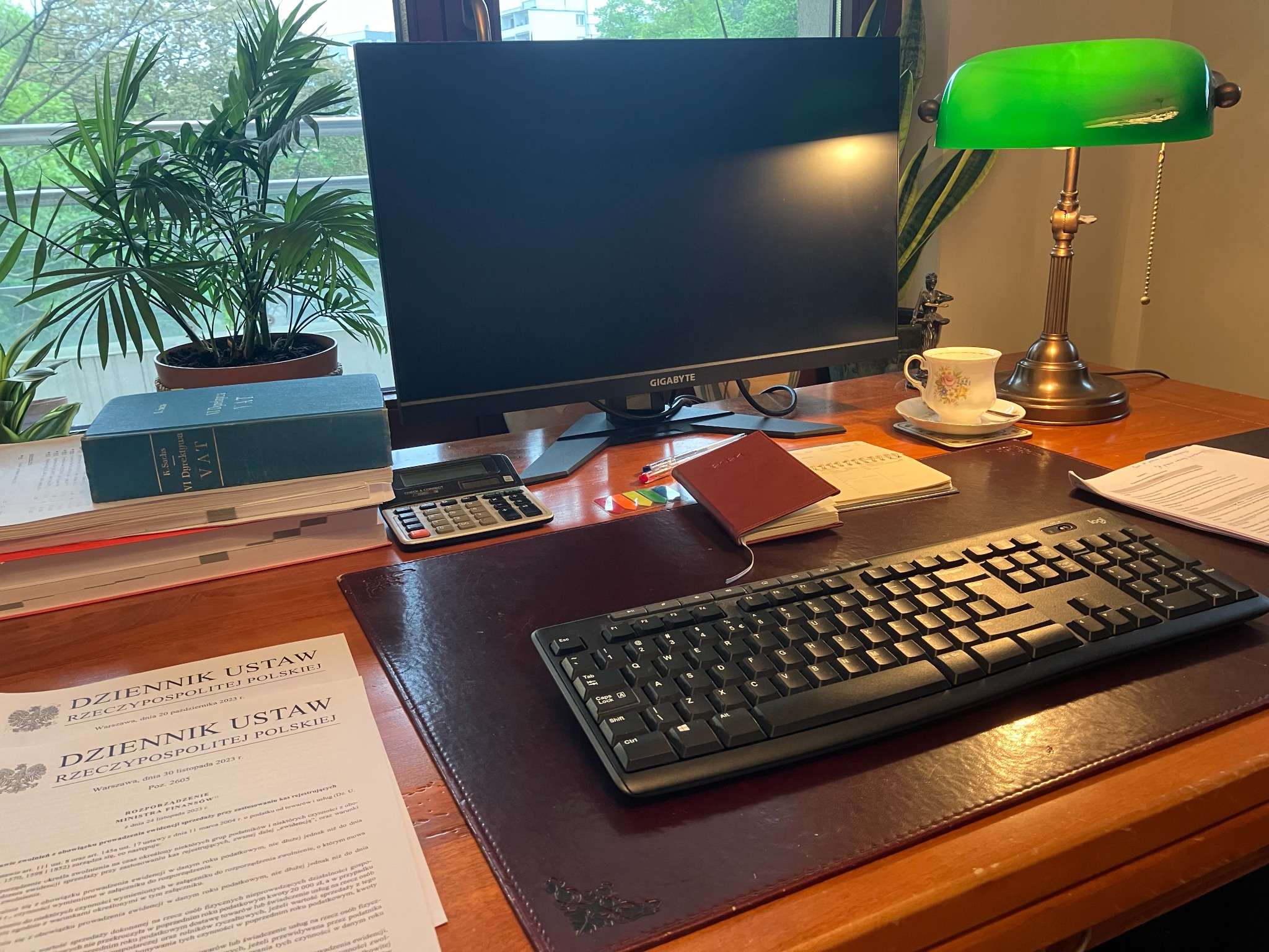 Biurko z dokumentami Dziennika Ustaw, kalkulatorem, klawiaturą, monitorem Gigabyte, lampką bankierską i rośliną doniczkową, sugerujące pracę w kancelarii.