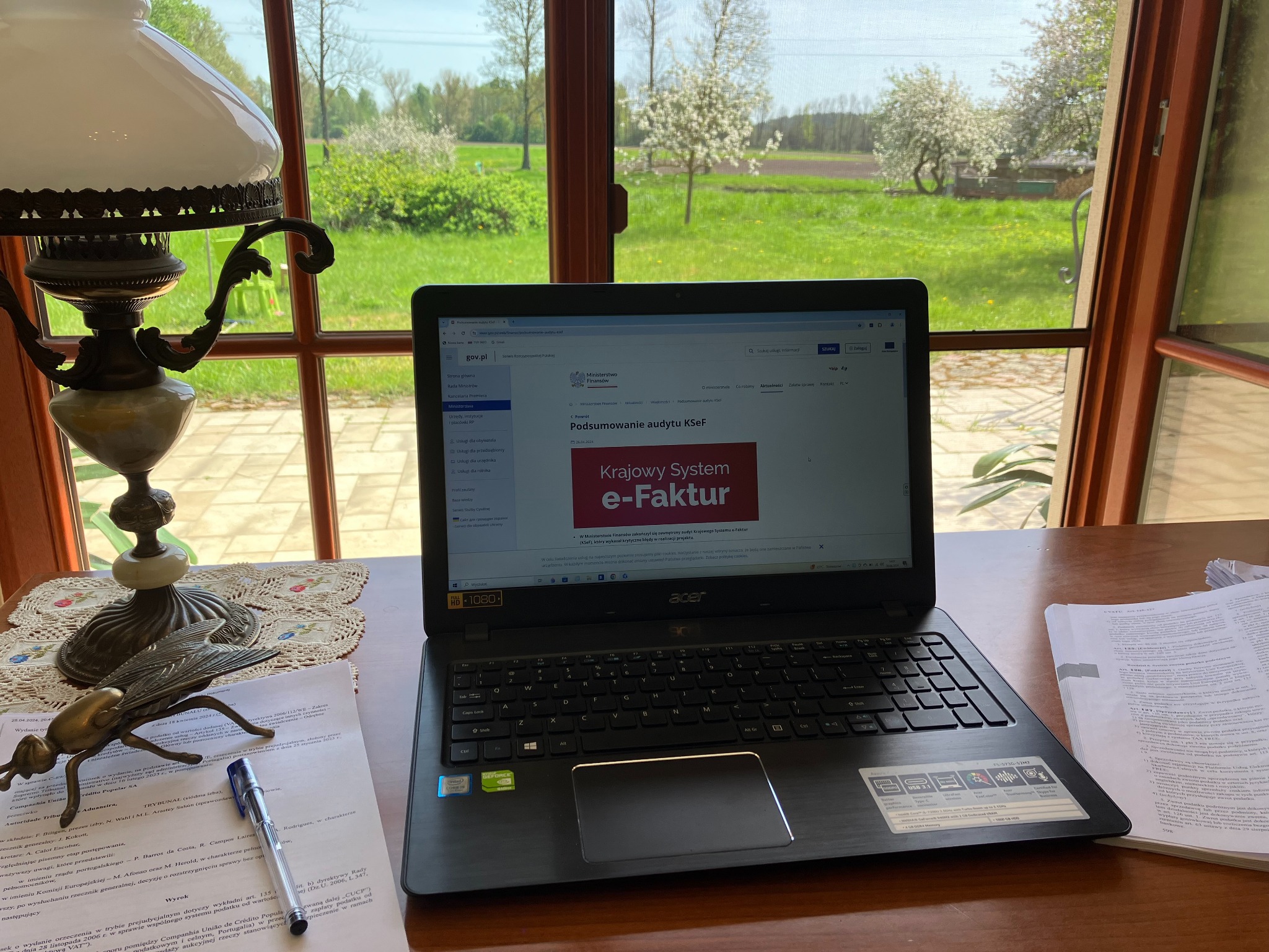 Biurko z laptopem wyświetlającym stronę Ministerstwa Finansów o Krajowym Systemie e-Faktur, obok lampa naftowa, metalowa figurka owada i dokumenty.