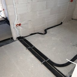 Marcin Polinarek - Surowe wnętrze w trakcie prac: kable elektryczne w czarnych peszlach, przymocowane metalowymi obejmami, prowadzone po szarej betonowej podłodze i ścianie z białych bloczków, widoczne puszki...