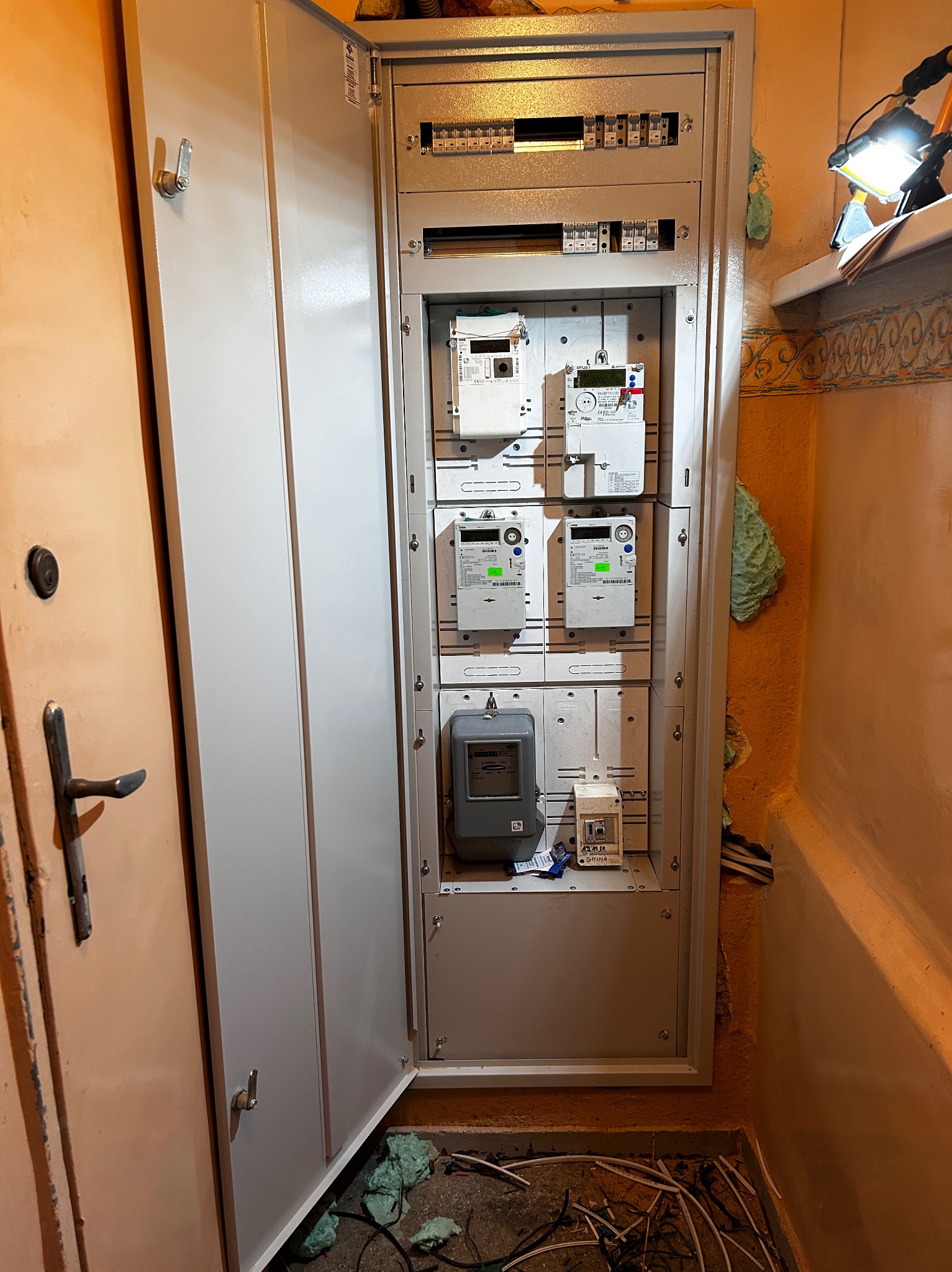 Otwarta szafka elektryczna z licznikami i bezpiecznikami, widoczne przewody na podłodze i lampa robocza w tle.