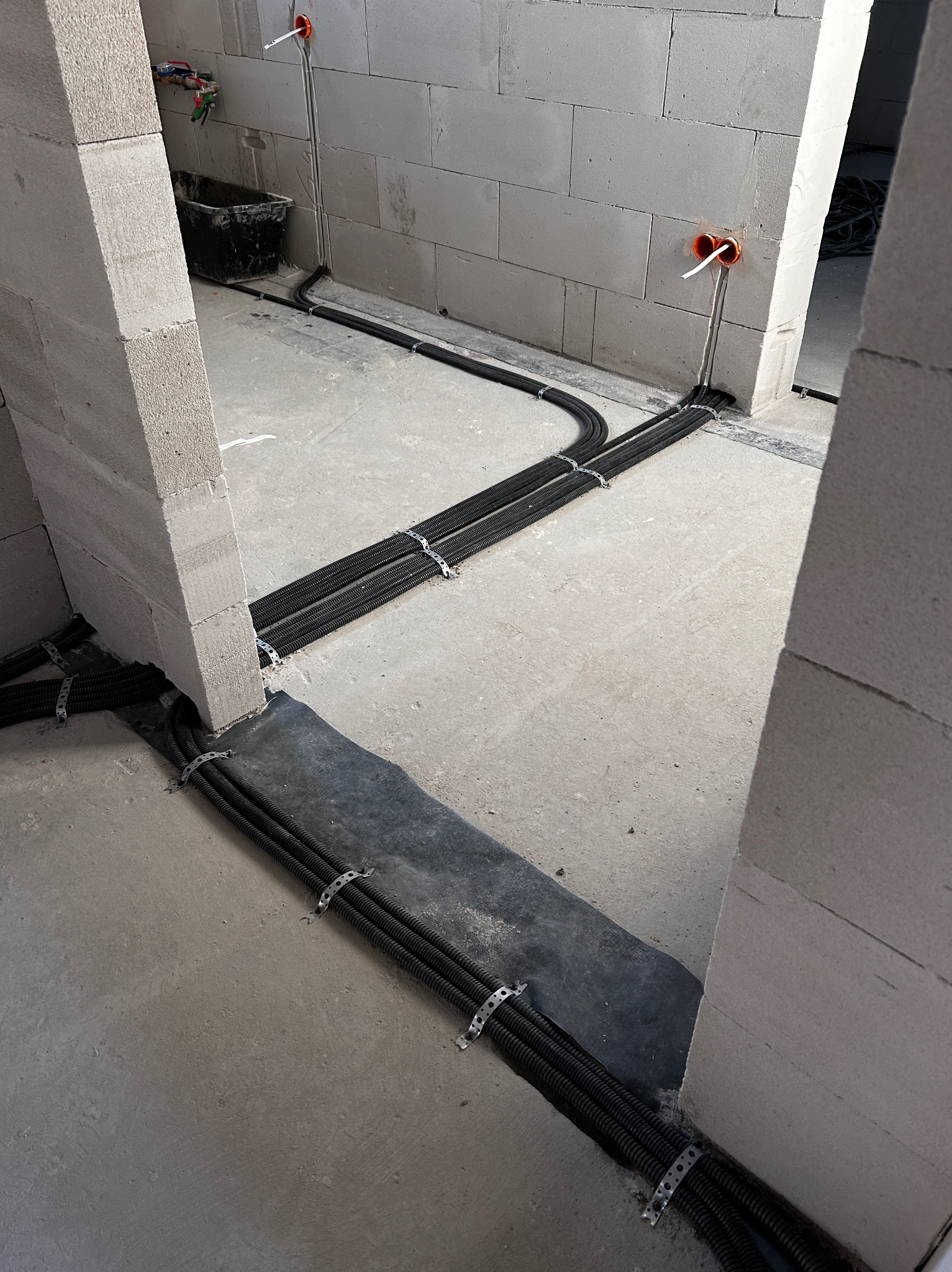 Surowe wnętrze z instalacją elektryczną w peszlach, przymocowaną metalowymi uchwytami do podłoża i ścian z betonu komórkowego. Widoczne puszki elektryczne w ścianach.