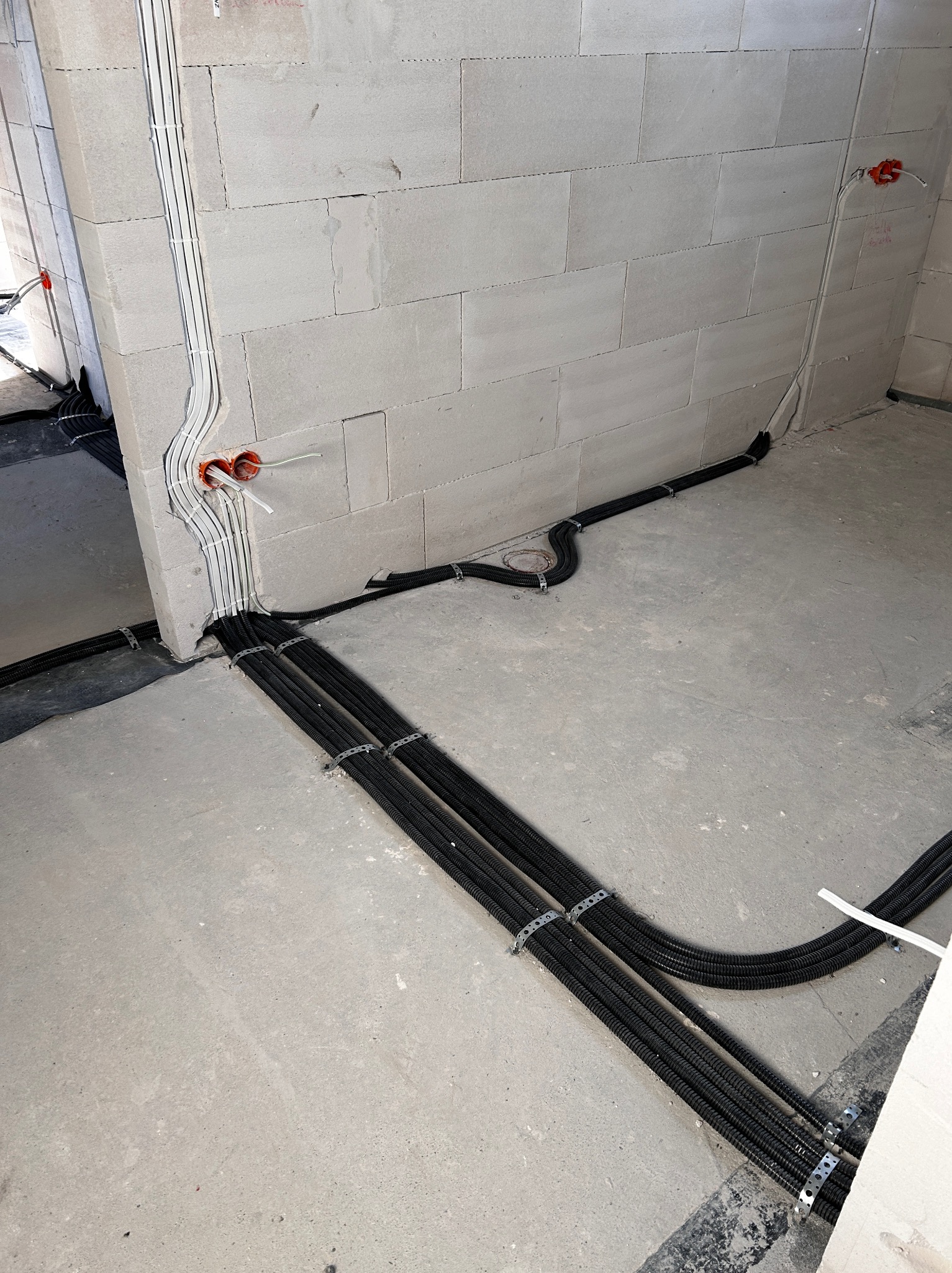 Surowe wnętrze w trakcie prac: kable elektryczne w czarnych peszlach, przymocowane metalowymi obejmami, prowadzone po szarej betonowej podłodze i ścianie z białych bloczków, widoczne puszki...