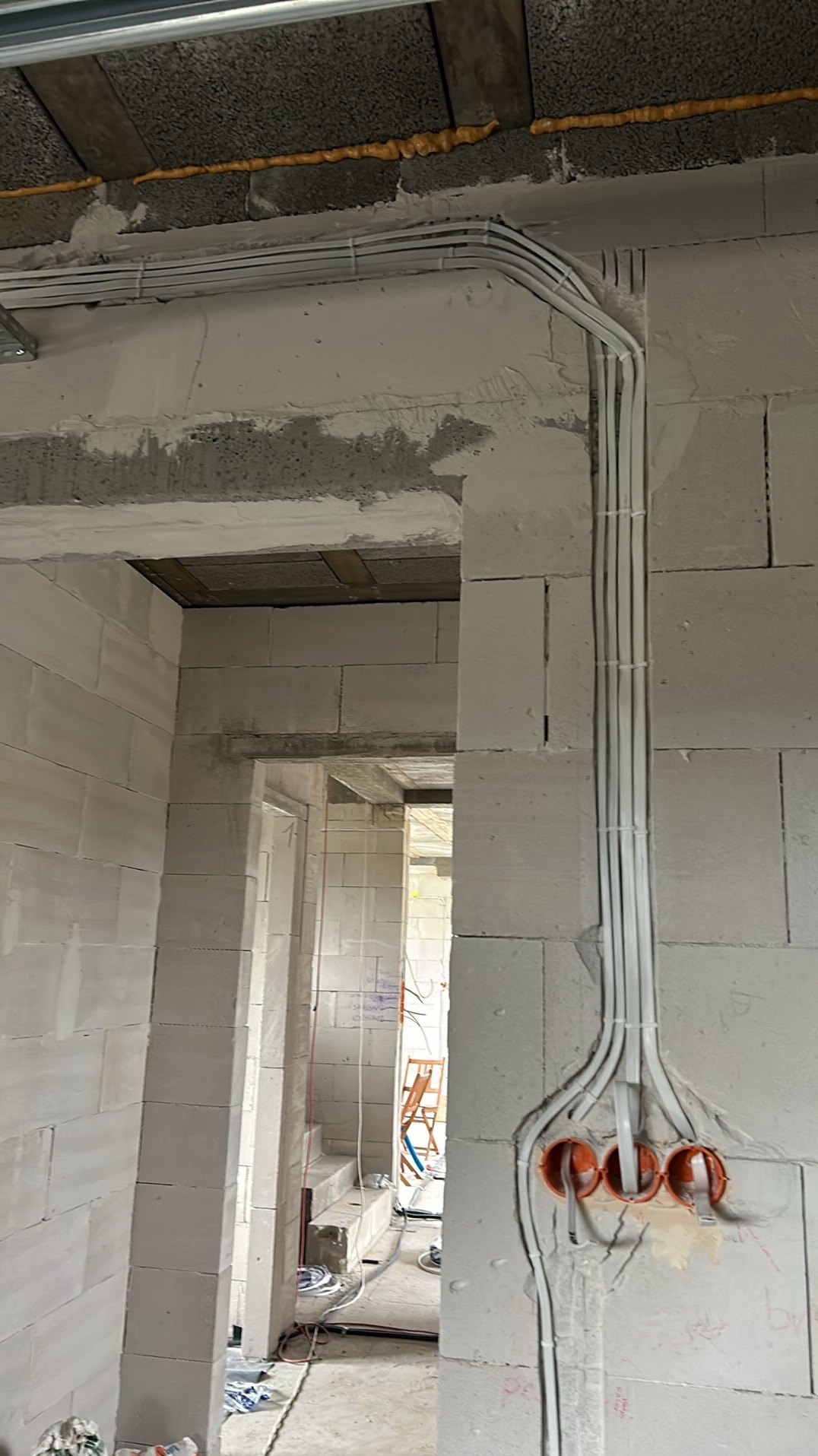 Wiązka przewodów elektrycznych w białej osłonie, przymocowana do ściany z betonu komórkowego za pomocą białych opasek zaciskowych, prowadząca do trzech pomarańczowych puszek instalacyjnych...