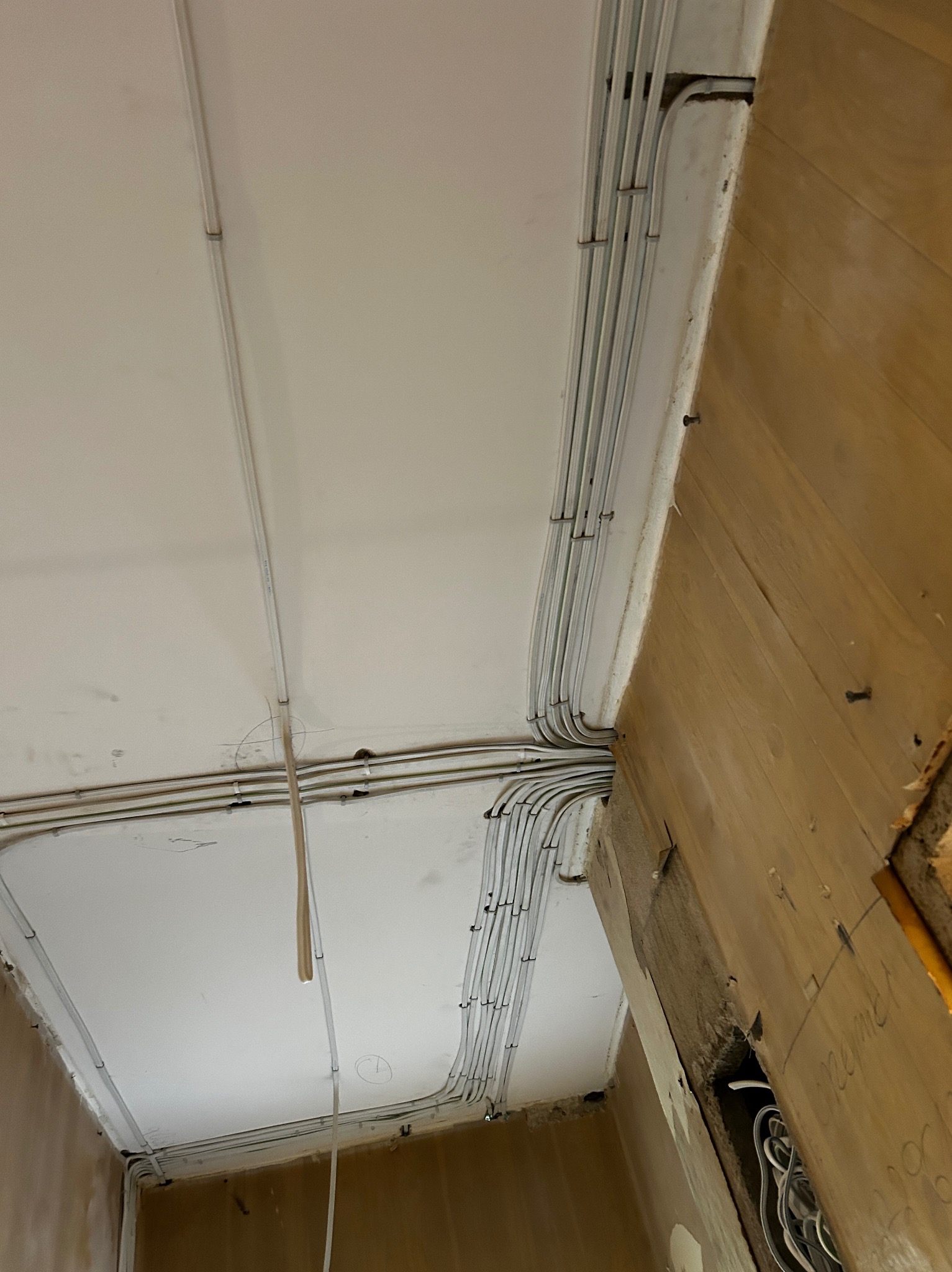 Biały sufit z widoczną instalacją elektryczną w rurkach PCV, prowadzoną wzdłuż krawędzi pomieszczenia, z widocznymi mocowaniami i fragmentem drewnianej ściany z instalacją wewnątrz.