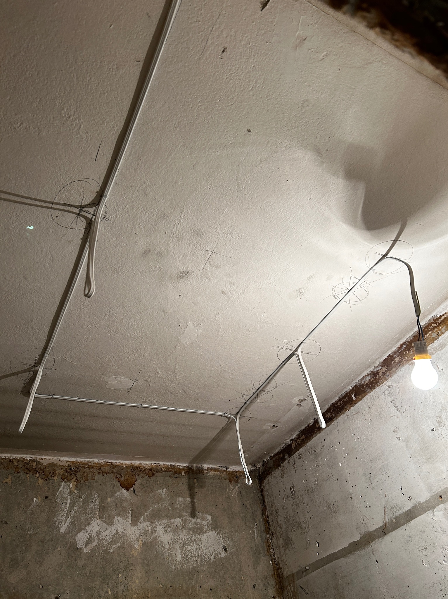 Surowy pokój w trakcie remontu z widoczną instalacją elektryczną natynkową, białe kable prowadzone po suficie i ścianach, wisząca żarówka oświetla wnętrze, ślady ołówka na suficie wskazują...