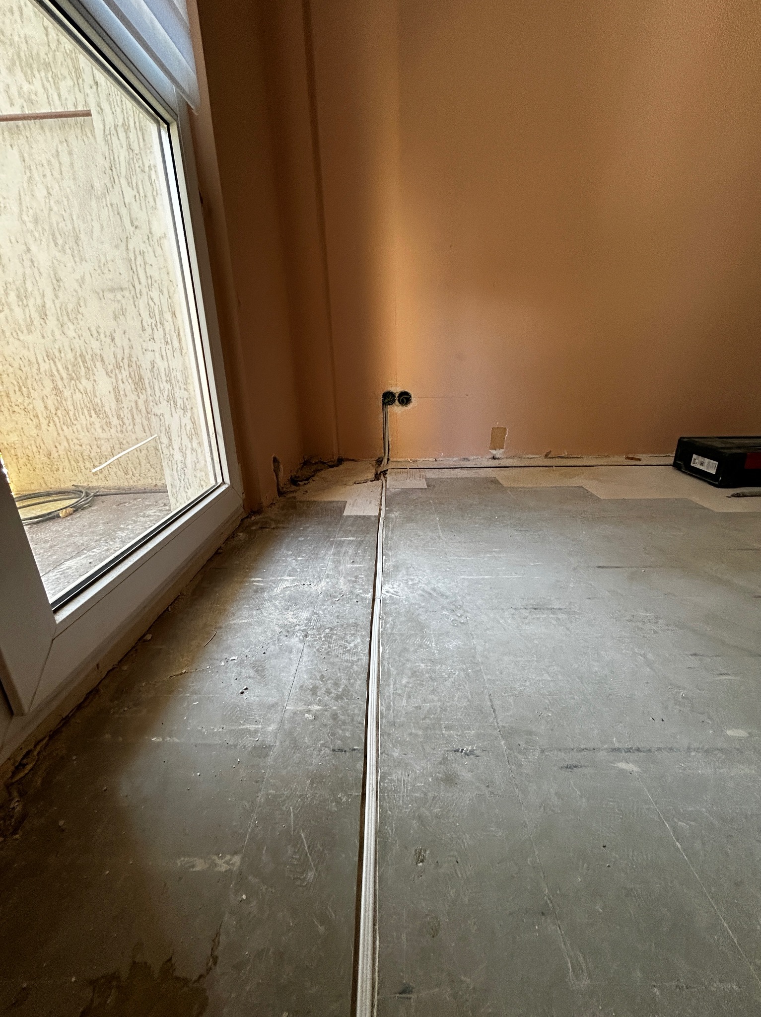 Wnętrze w trakcie remontu w Wolsztynie: widoczne gniazdka elektryczne w ścianie, ślady po przewodach w podłodze i surowy beton. Przy oknie leżą kable.