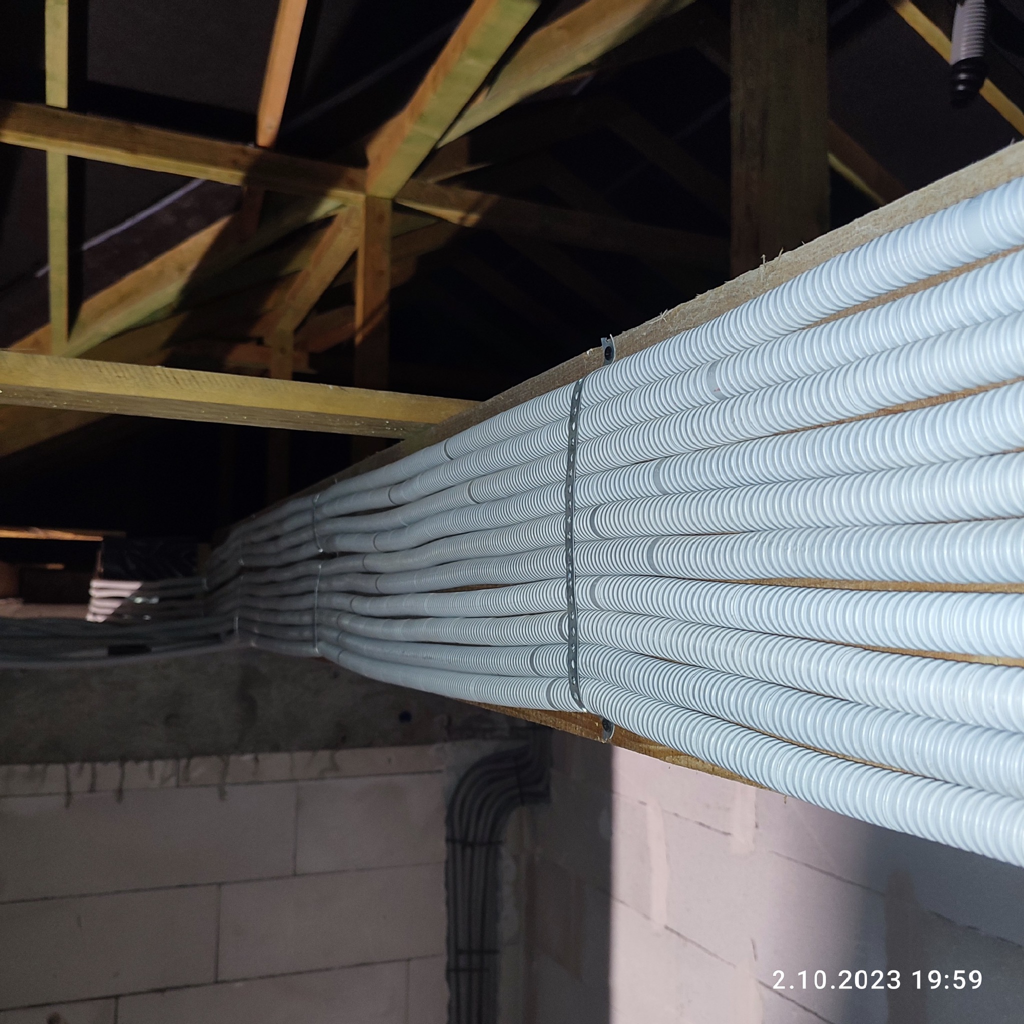 Równolegle ułożone, szare rury karbowane przymocowane do drewnianej belki stropowej w surowym wnętrzu budynku z betonowych pustaków.
