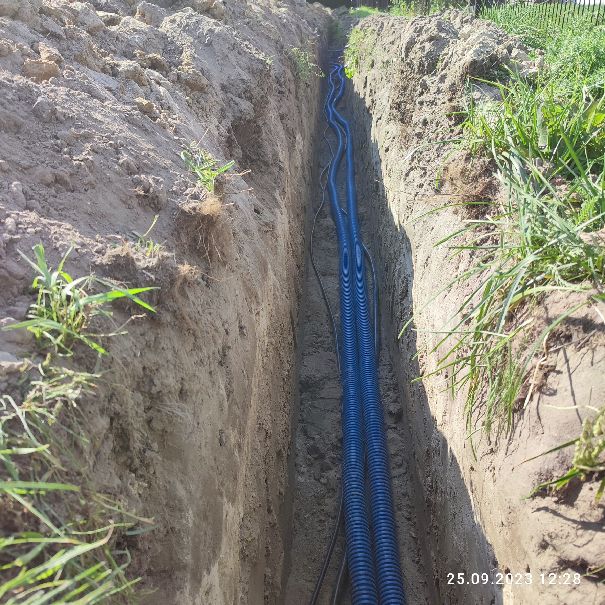 Wykop w ziemi z ułożonymi niebieskimi rurami karbowanymi i czarnymi kablami, widok z góry.