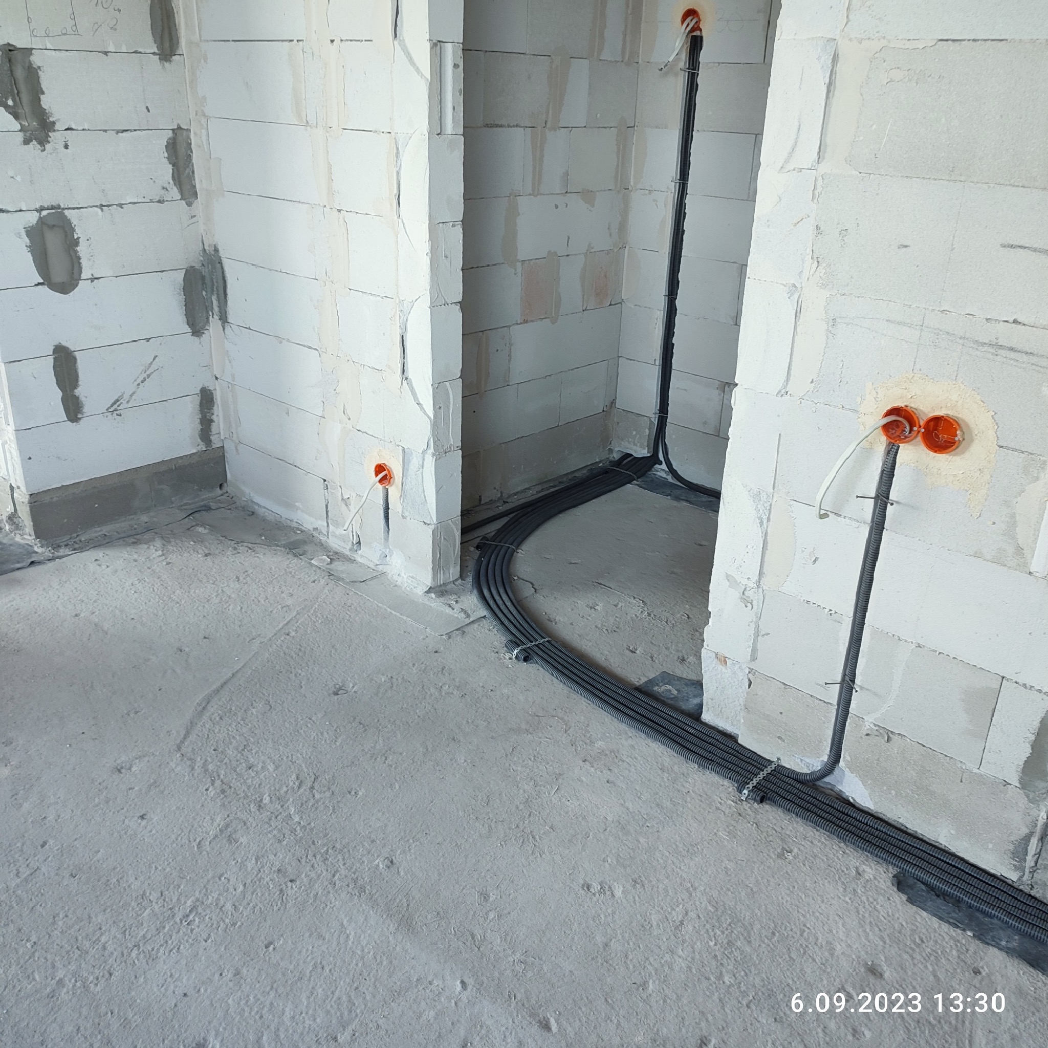 Surowy stan: Kable elektryczne w peszlach ułożone na podłodze w trakcie prac instalacyjnych w budynku z białych bloczków, widoczne puszki elektryczne w ścianach.