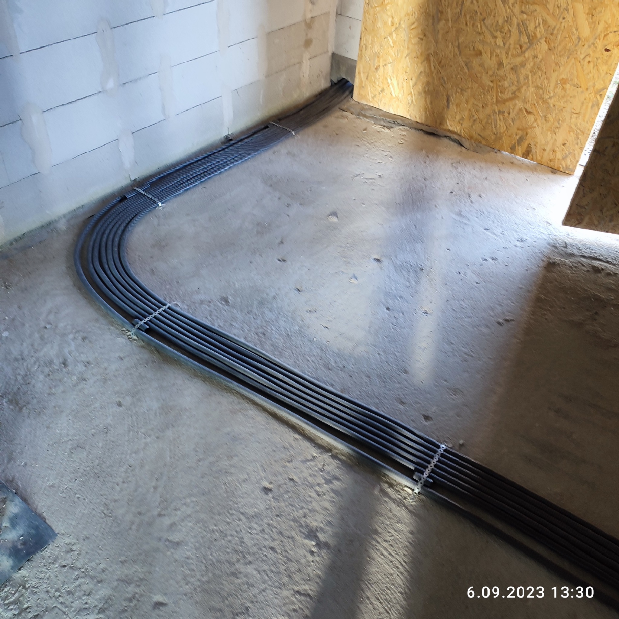 Wiązka szarych rur karbowanych ułożona na surowym betonowym podłożu wzdłuż ściany z bloczków betonowych, zabezpieczona spinkami, prowadząca do otworu w ścianie z płyty OSB.