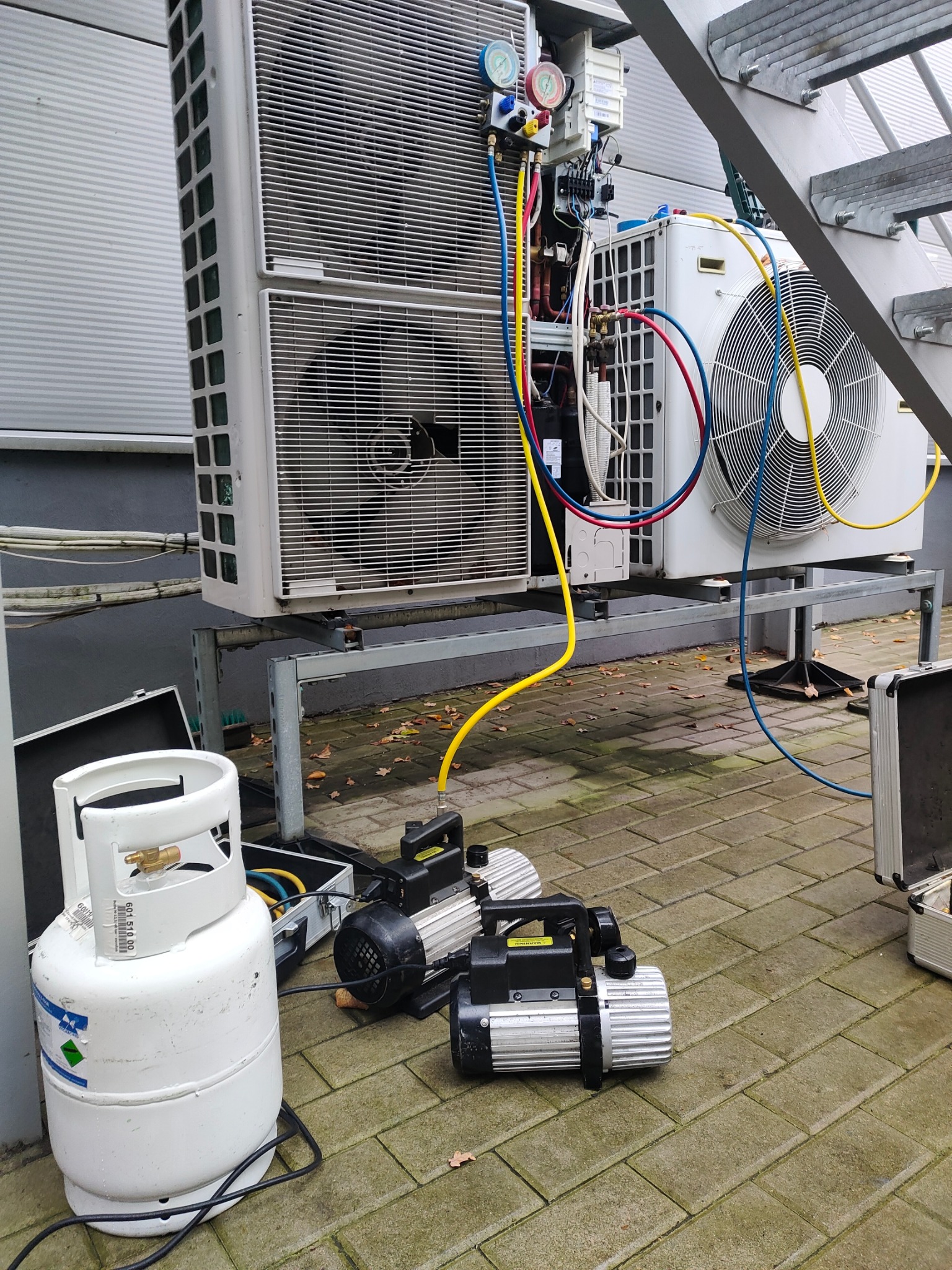 Dwa agregaty klimatyzacyjne na metalowej konstrukcji, podłączone przewodami do manometrów, obok butla z czynnikiem chłodniczym i dwie pompy próżniowe na kostce brukowej, w tle schody stalowe.