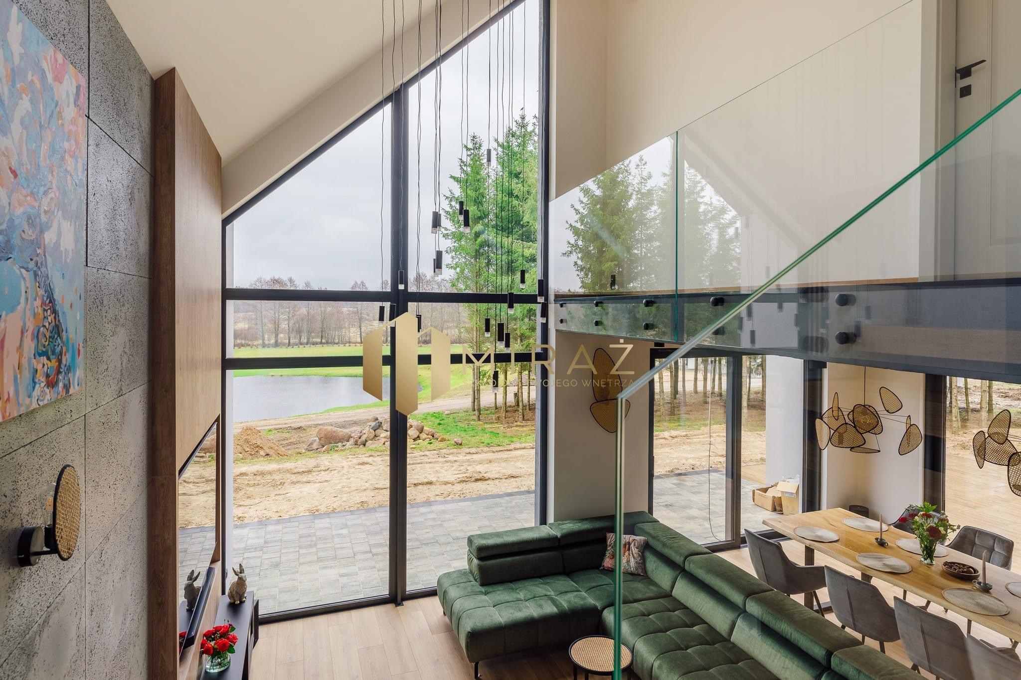 Wnętrze nowoczesnego domu z dużymi oknami, zieloną sofą modułową, szklaną balustradą na antresoli i widokiem na jezioro. W tle widoczny teren budowy.