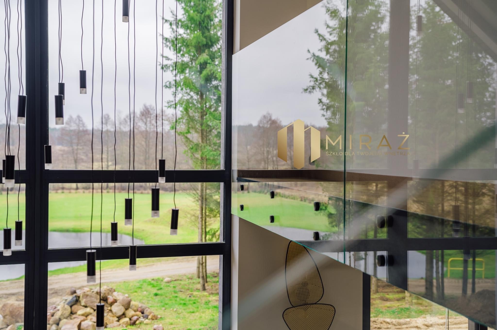 Szklana balustrada schodowa z widocznym logo firmy 'Miraż Szkło dla Twojego Wnętrza', czarne, metalowe elementy konstrukcyjne, tło stanowi widok na zewnątrz przez okno z wiszącymi lampami.