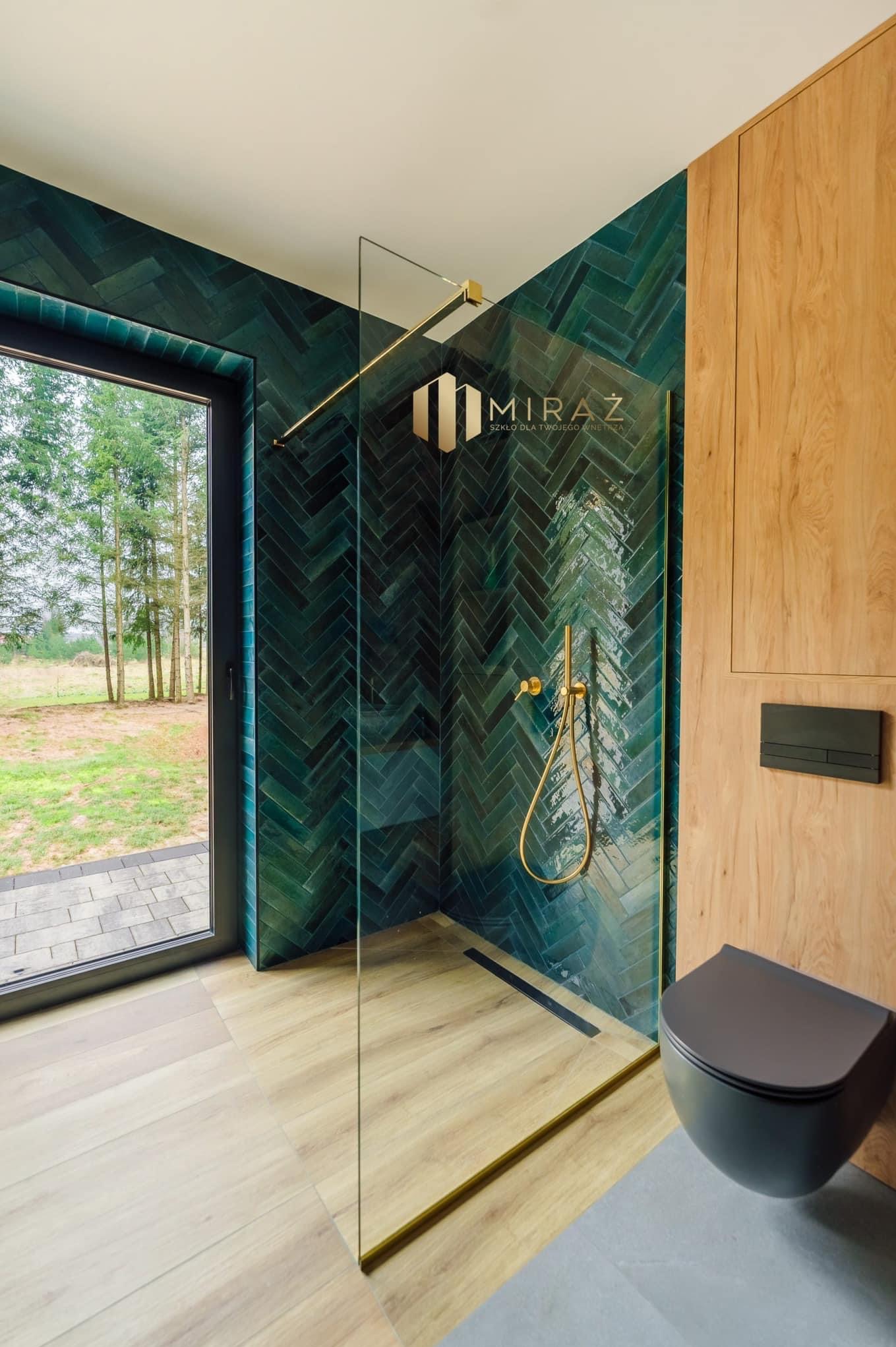 Nowoczesna łazienka z kabiną prysznicową ze szkła, ciemnozielone płytki ułożone w jodełkę, złota armatura i drewniane panele ścienne, widok na zewnątrz przez czarne okno.