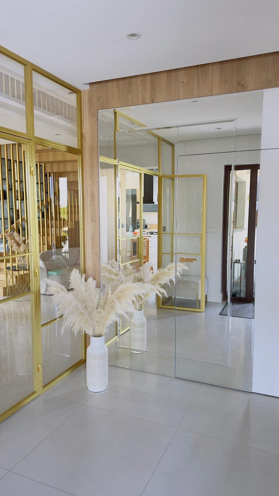 Nowoczesne wnętrze z lustrzaną ścianą w złotej ramie, dekorowane białymi wazonami z pampasową trawą, widok na salon i kuchnię przez szklane drzwi.