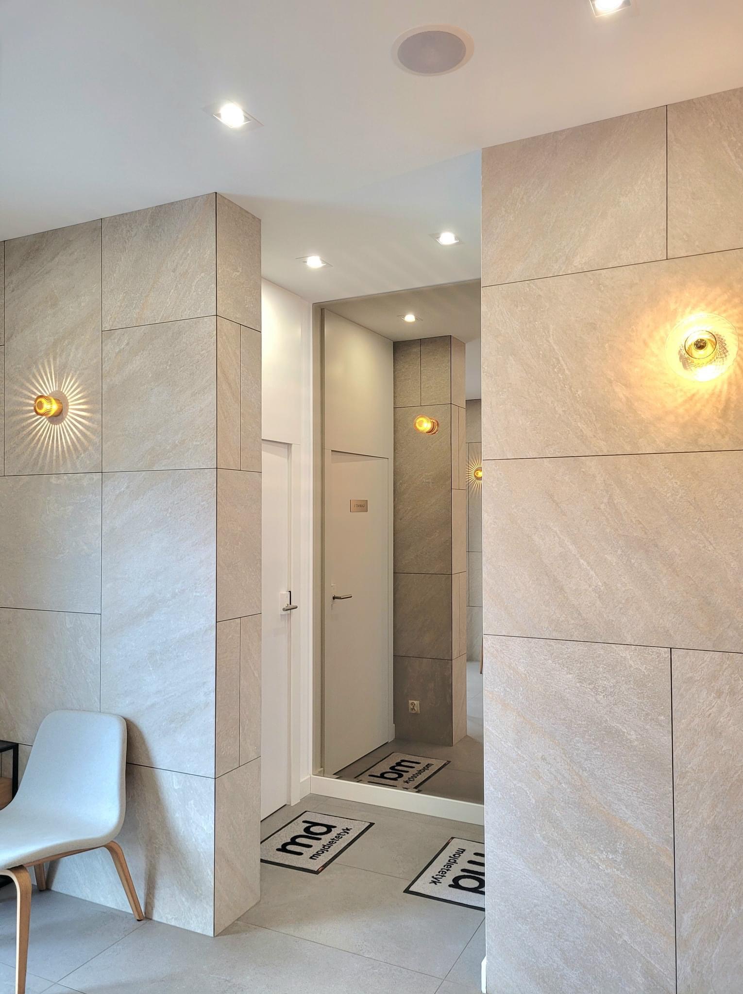 Hall z płytkami imitującymi kamień w odcieniach beżu, oświetlony dekoracyjnymi kinkietami i lampami sufitowymi. W lustrze odbicie dalszej części korytarza z widocznymi wycieraczkami z logo.