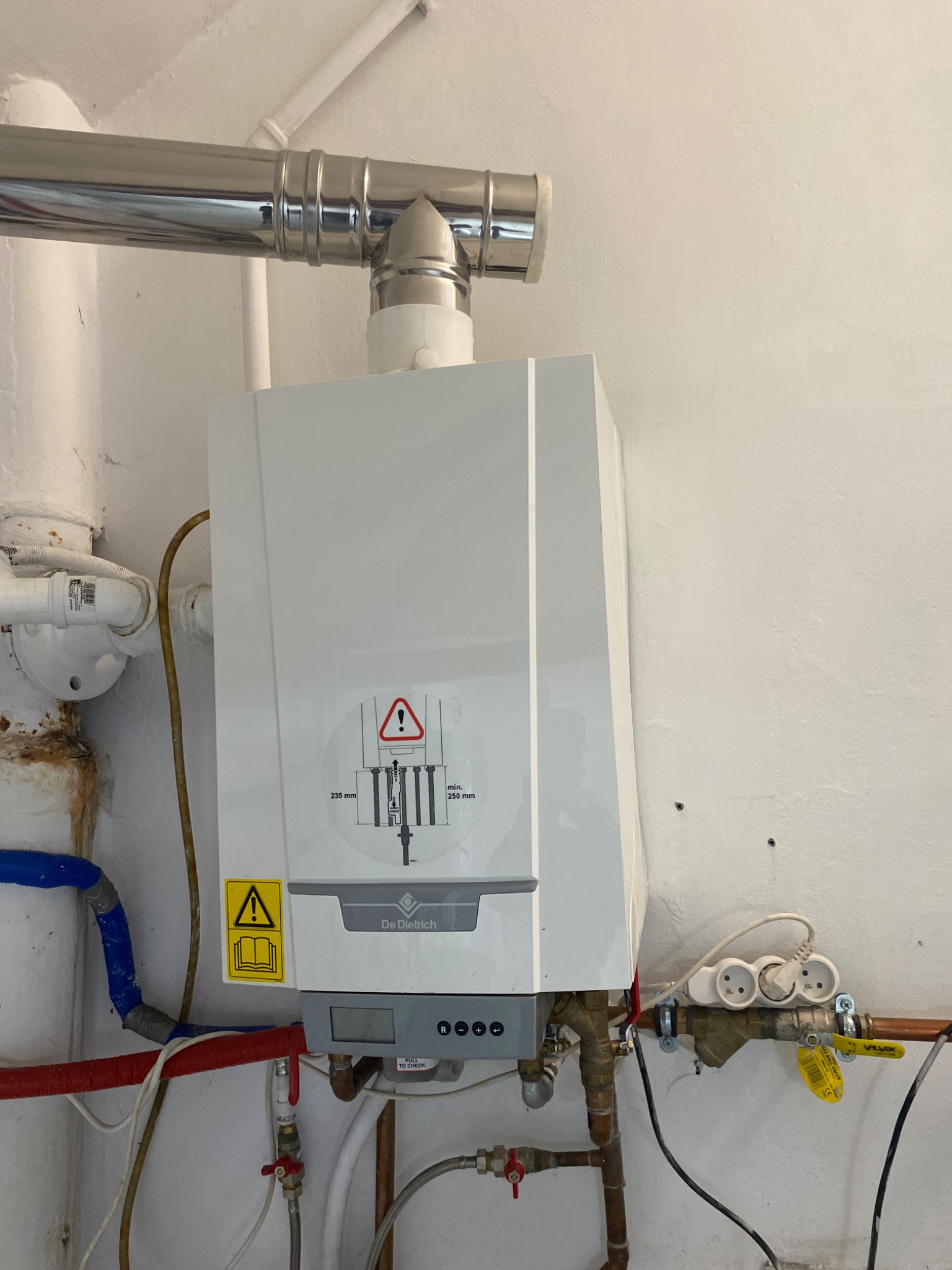 Kocioł grzewczy De Dietrich zamontowany na ścianie, z widocznymi rurami doprowadzającymi i odprowadzającymi wodę oraz spalinami, gniazdko elektryczne w pobliżu.
