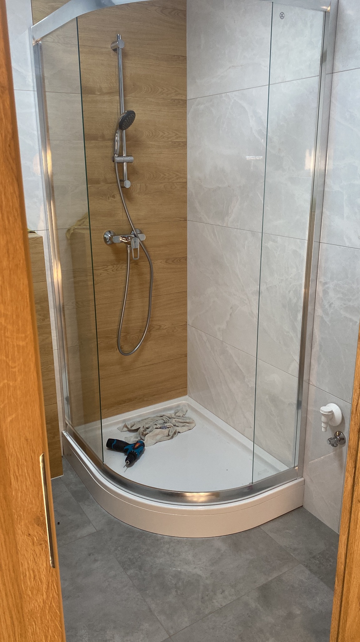 Nowa kabina prysznicowa z chromowaną armaturą, zamontowana na białym brodziku, z widoczną wkrętarką i szmatką wewnątrz, na tle płytek imitujących drewno i marmur.