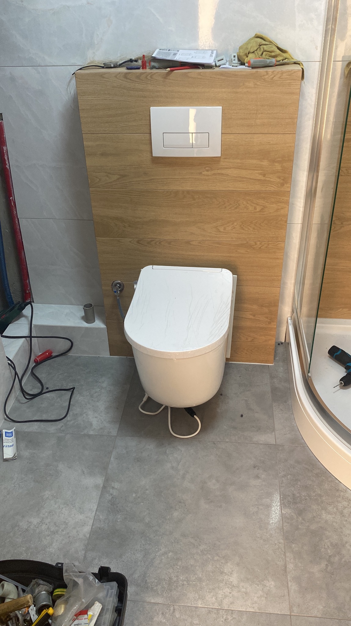 Montaż wiszącej toalety myjącej z podtynkową spłuczką na tle płytek imitujących drewno, widoczne narzędzia i elementy instalacji.