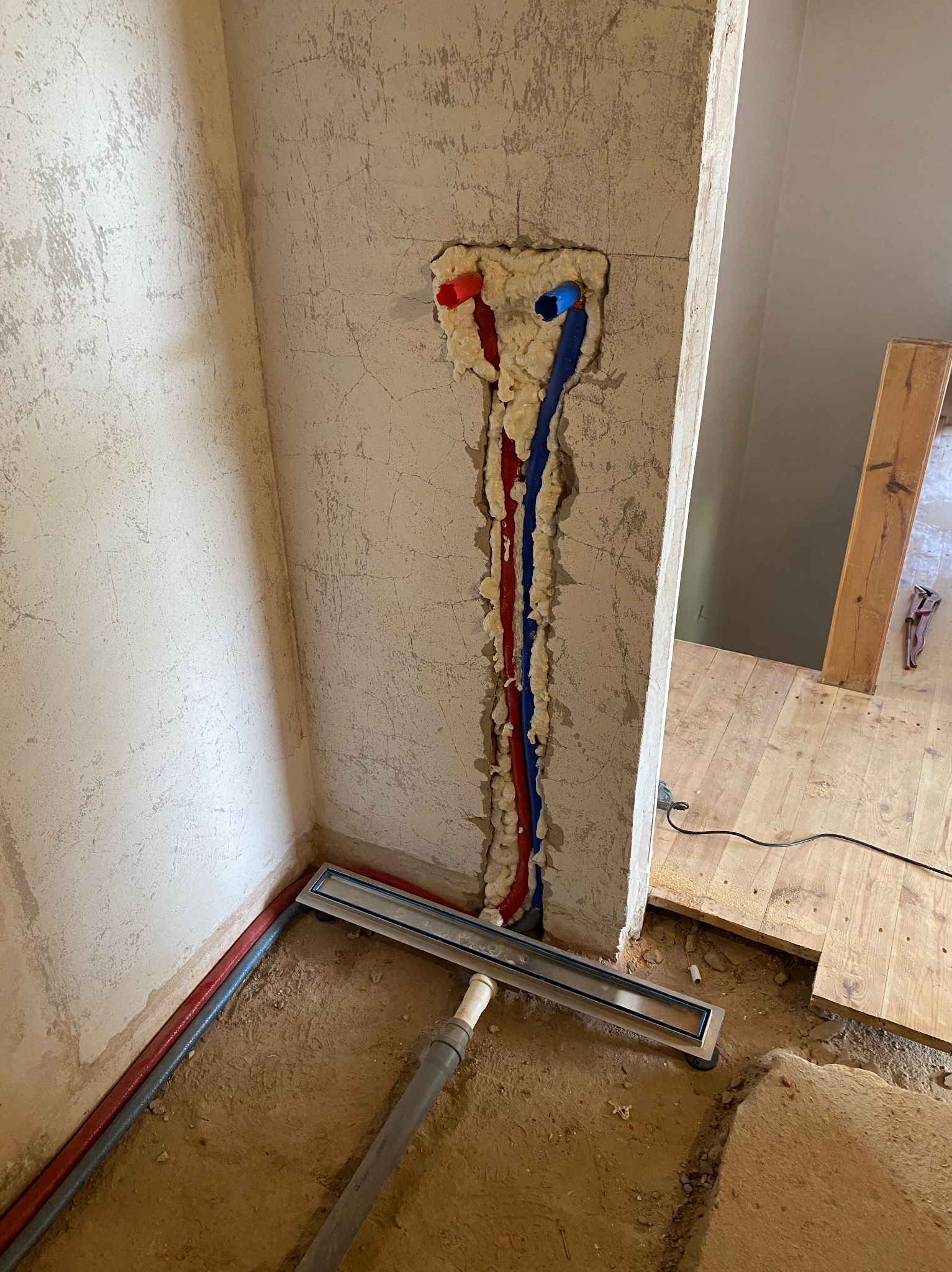Fragment łazienki w trakcie remontu: widoczne rury (czerwona i niebieska) osadzone w ścianie pianką montażową, odpływ liniowy w podłodze, surowe ściany z widocznymi śladami po poprzednich płytkach.