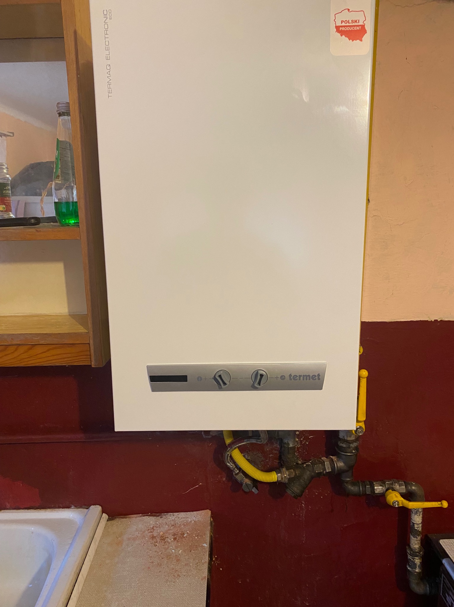 Wiszący, biały, gazowy podgrzewacz wody marki Termaq Electronic Eco z naklejką 'Polski Producent', podłączony do instalacji z żółtymi rurami gazowymi i zaworem, na tle czerwonej ściany i fragmentu...