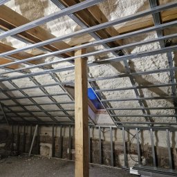 Marci Marcin - Poddasze w trakcie ocieplania pianką PUR, widoczne krokwie, metalowy stelaż pod płyty gipsowo-kartonowe i częściowo nałożona pianka, z niebieskim oknem dachowym w tle i drewnianym słupem...