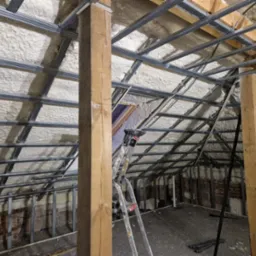 Poddasze w trakcie prac: ocieplenie pianką PUR na skośnym dachu, widoczne metalowe profile konstrukcyjne, drewniane słupy podtrzymujące, drabina i odsłonięta cegła na ścianach