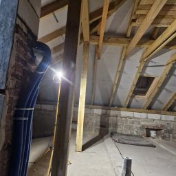 Marci Marcin - Poddasze w trakcie remontu, widoczne drewniane krokwie, izolacja, niebieskie rury wentylacyjne mocowane do ściany z cegły, oświetlenie robocze
